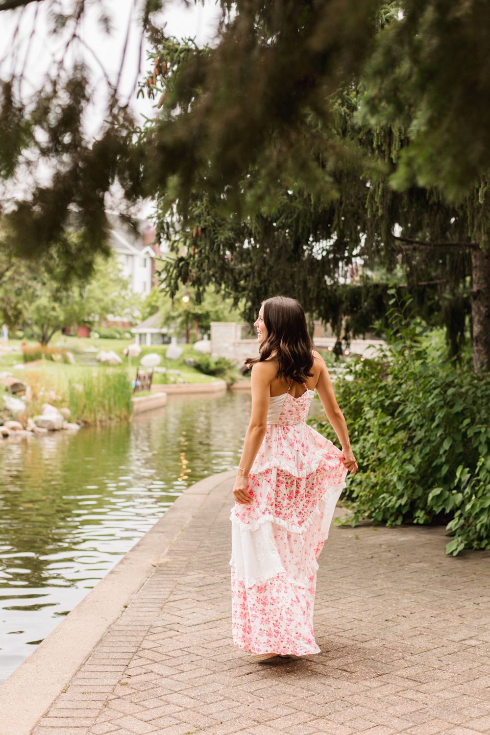 A woman in a flowy red and white dress spins gracefully along a lakeside brick path surrounded by greenery.