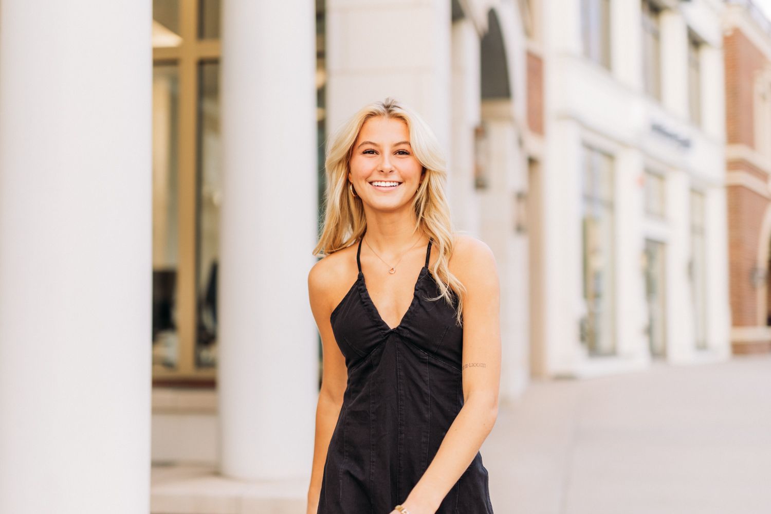 A person in a black sleeveless top poses confidently in an outdoor setting with white columns and archways.