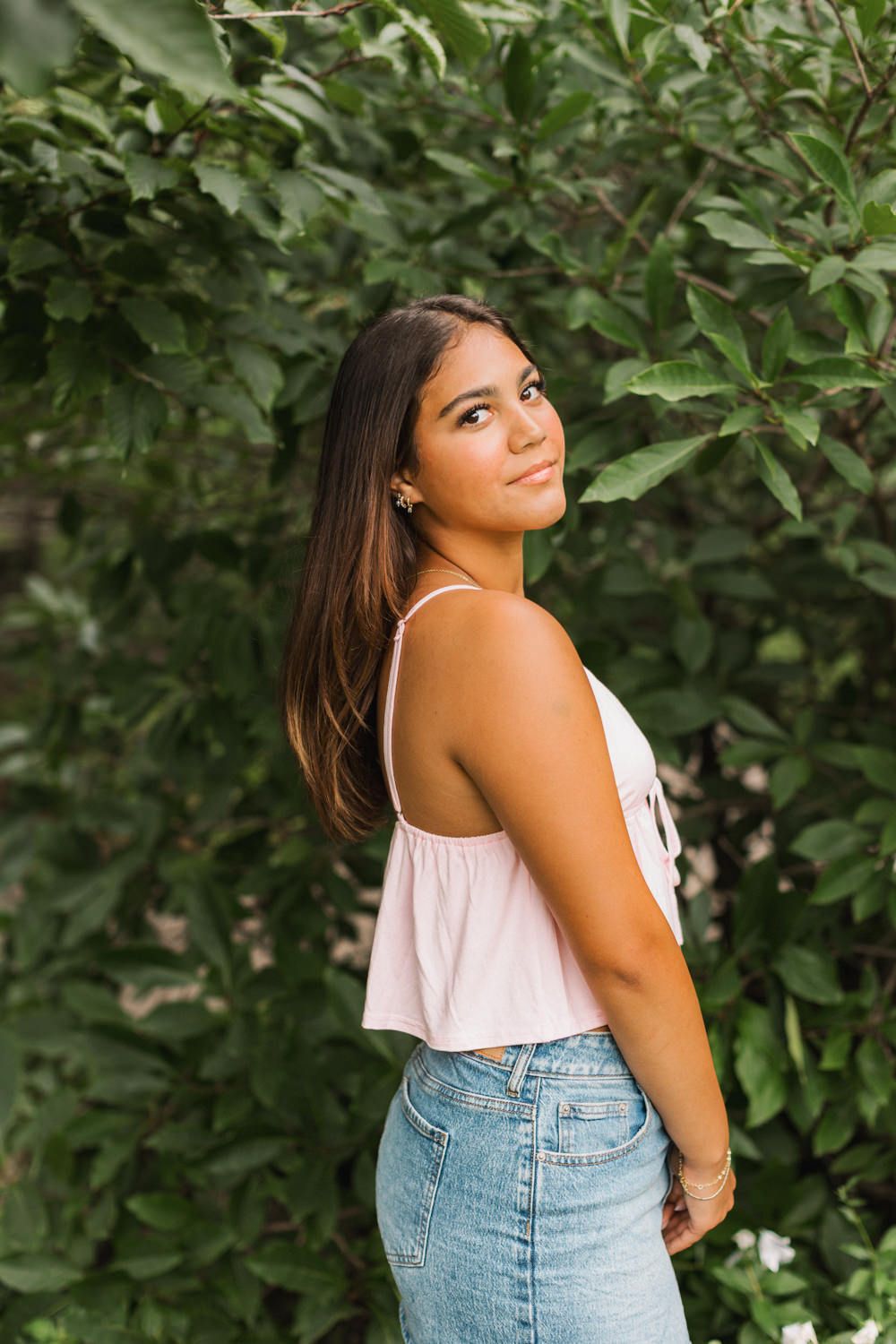 Side profile portrait showing a pink top and denim outfit against lush green foliage.