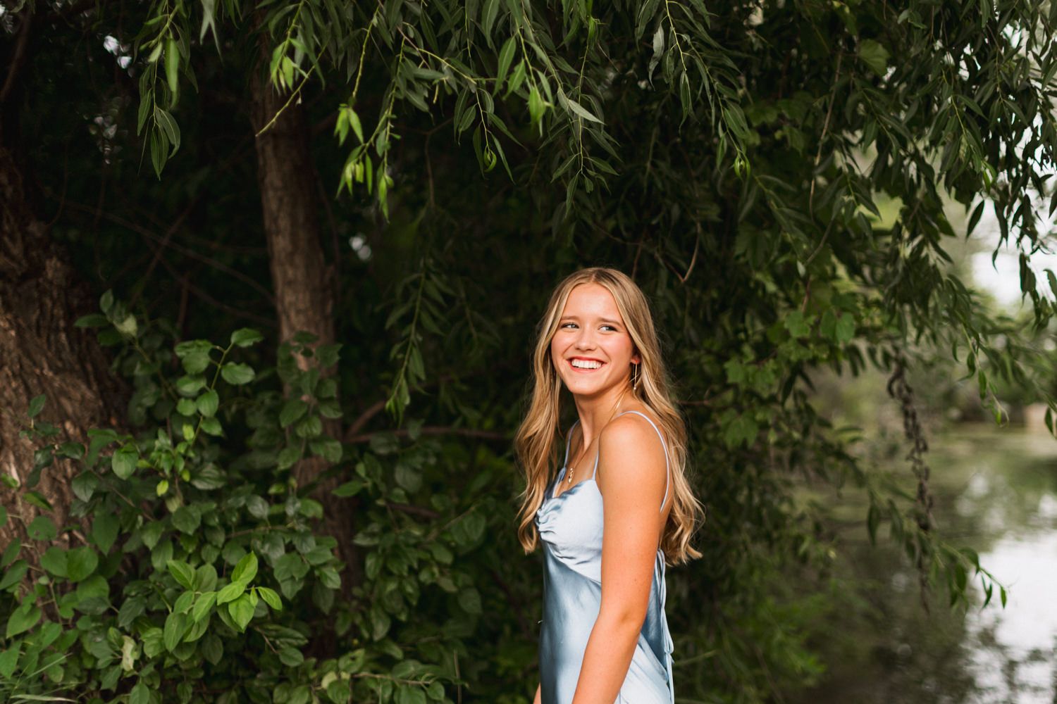 Someone in a light blue dress poses under hanging willow tree branches near water.
