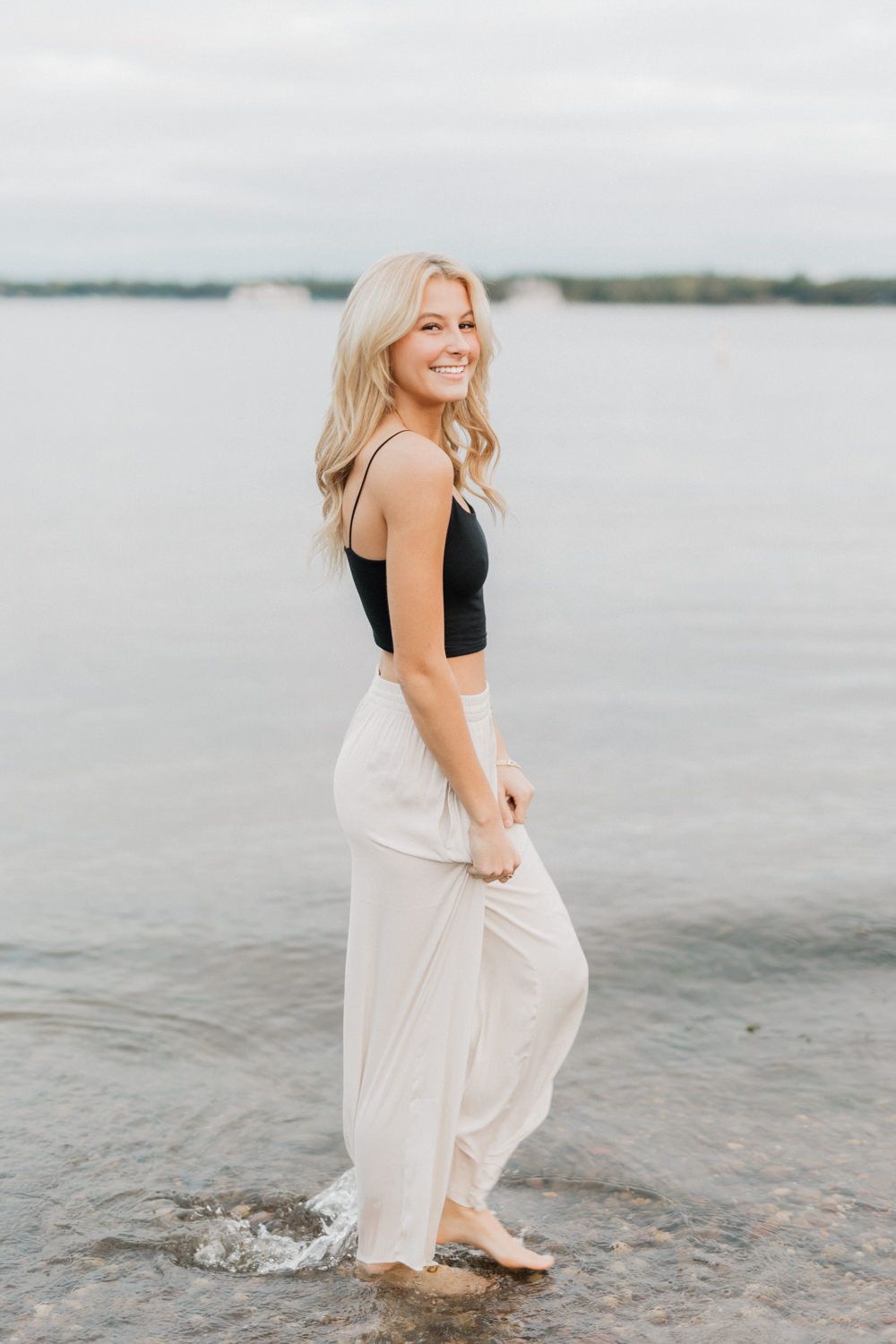 A person in a black top and flowing white skirt playfully splashes in shallow coastal waters at sunset.