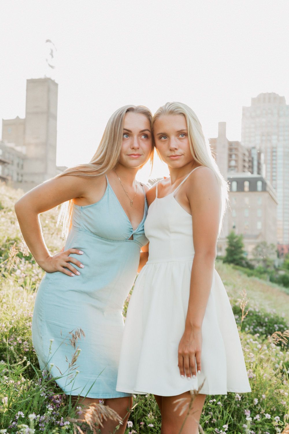 Two women in summer dresses pose together in a grassy field with city buildings in the background.