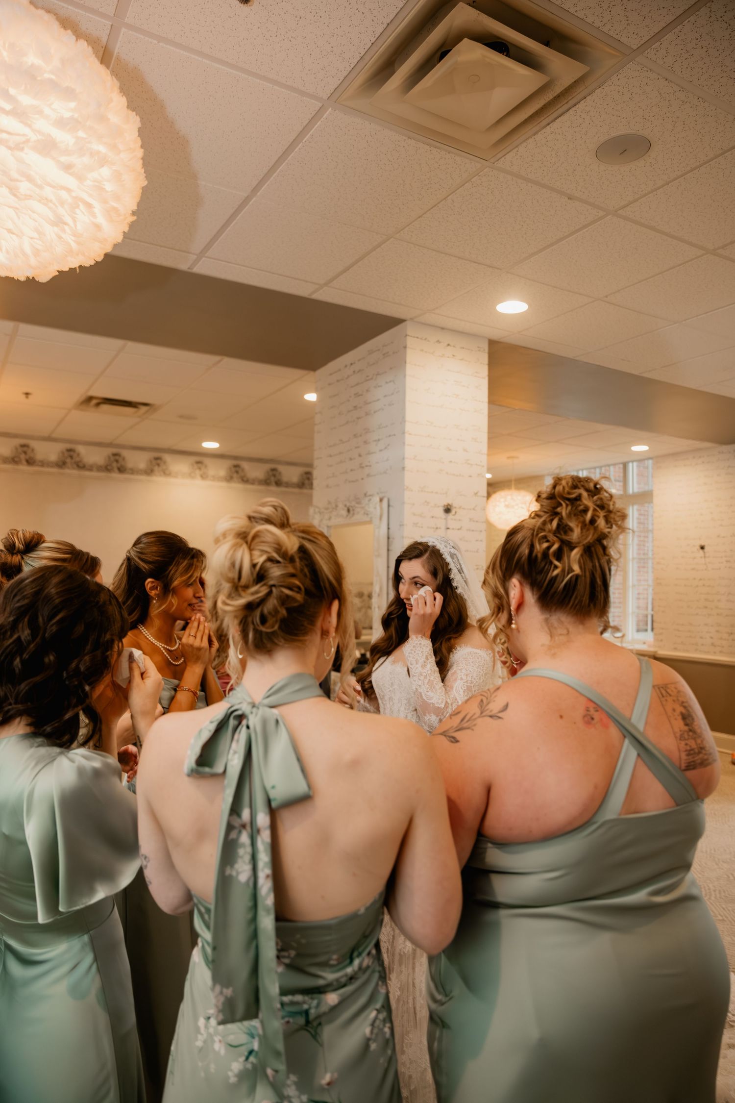 Bridesmaids in sage green dresses gather around an emotional bride in a white robe during wedding preparations.