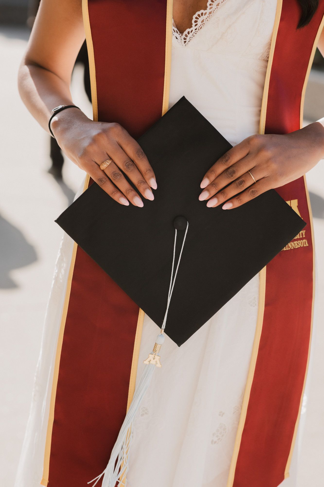 Close-up of graduate holding black mortarboard cap with tassel while wearing maroon and gold graduation stole.