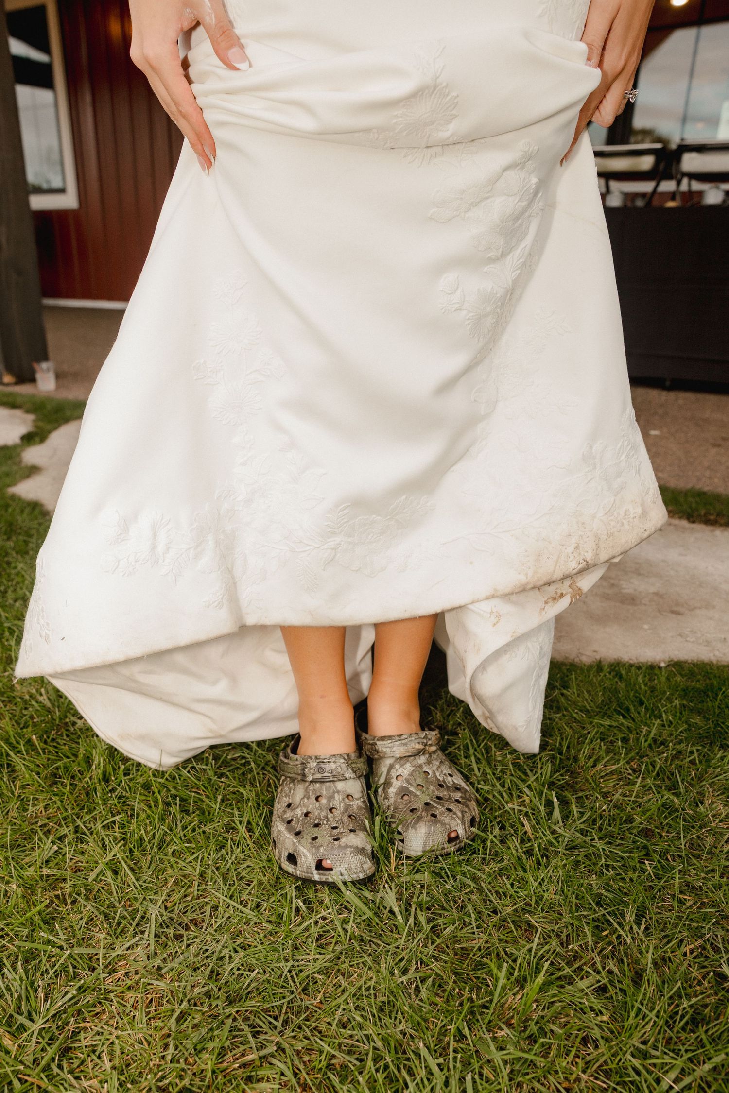 Bride lifts white wedding gown to reveal muddy Crocs shoes while standing on green grass outdoors.