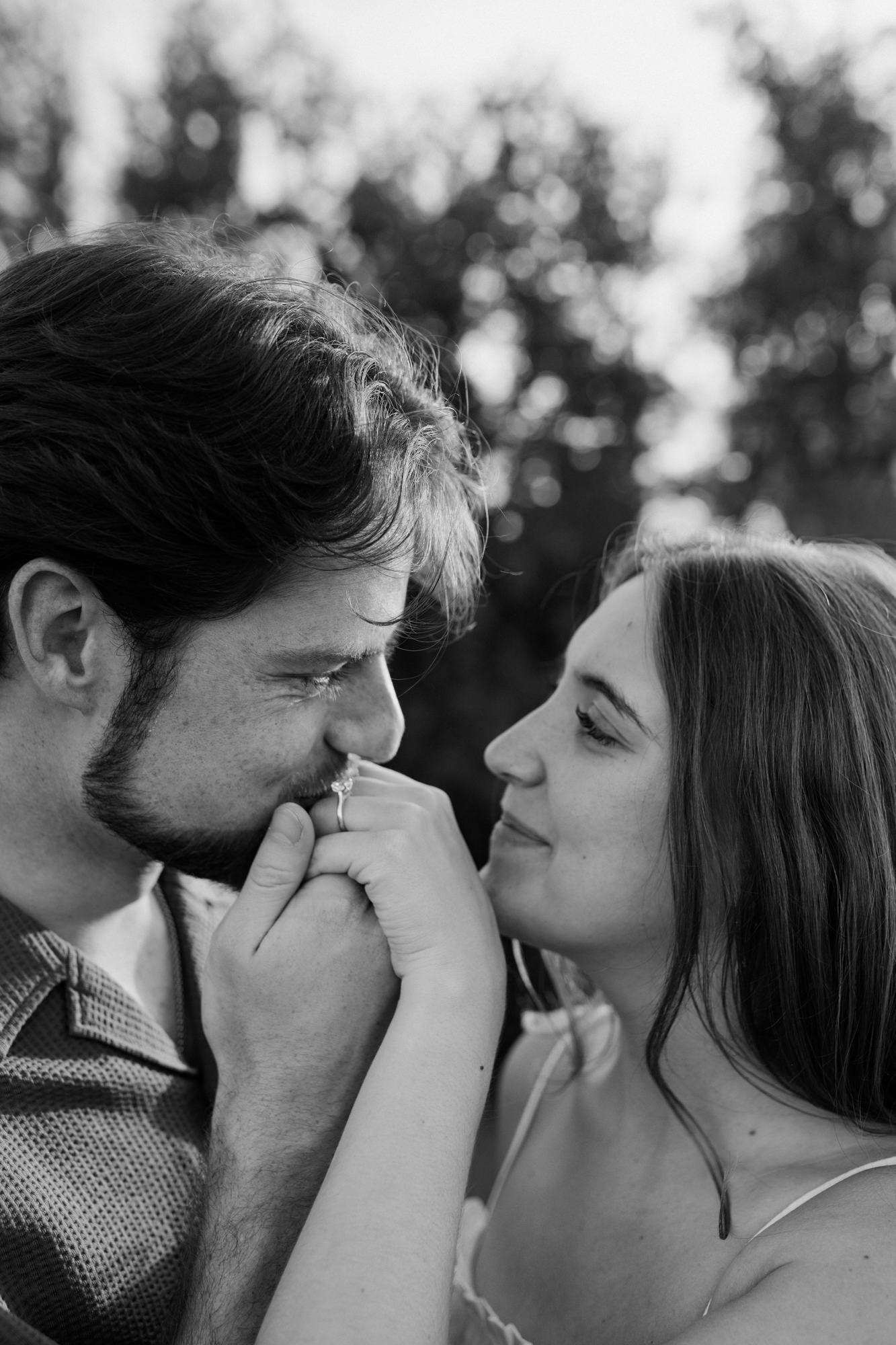 Black and white photo of a romantic couple holding hands and gazing at each other outdoors with trees in the background.