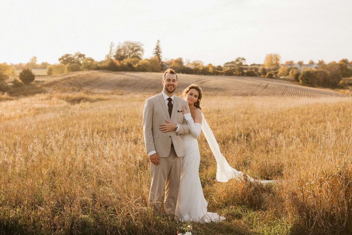 Bride and groom in a tan suit embrace in a golden grass field at sunset, her veil trailing in the breeze.