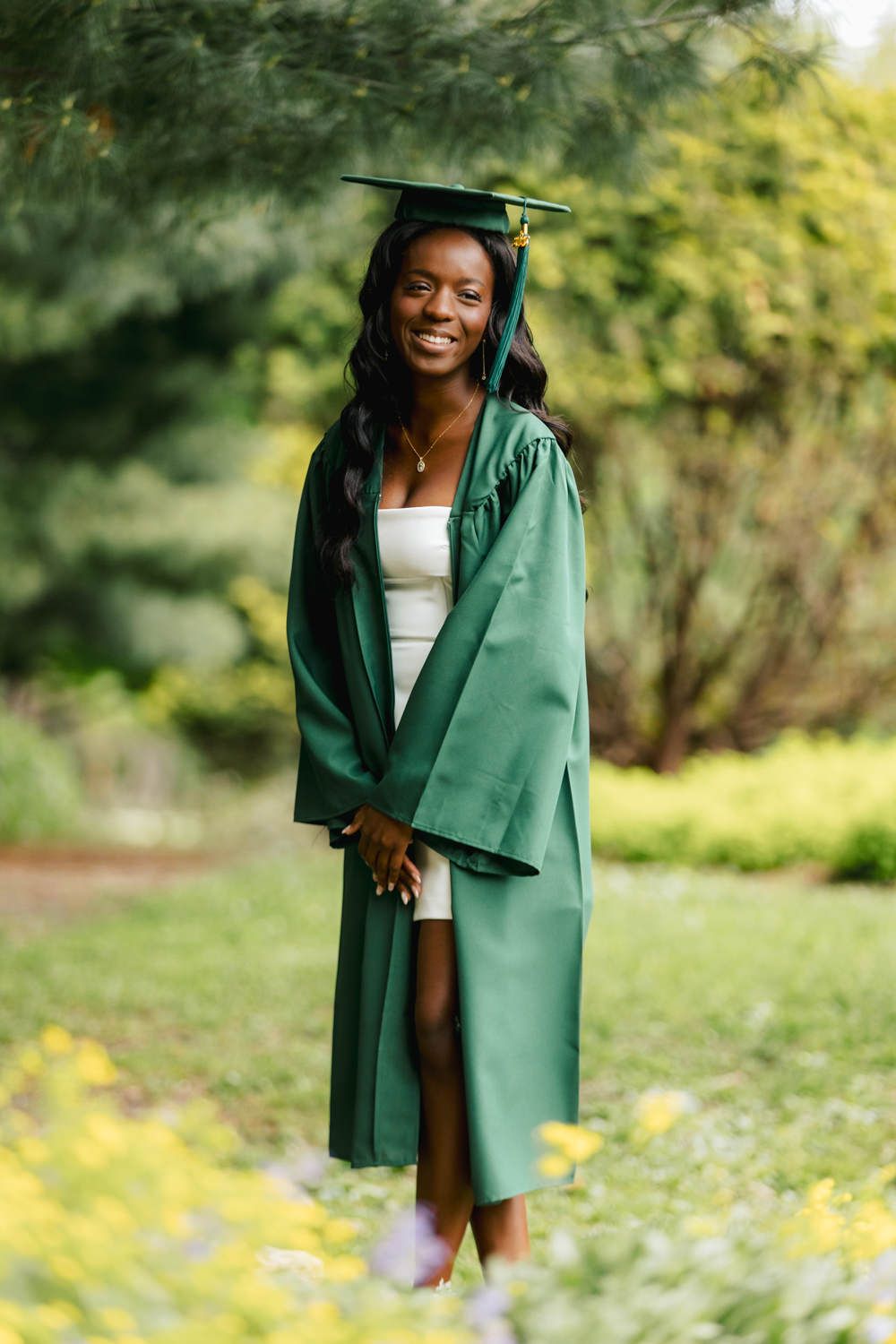 Graduate in flowing green graduation gown poses against blurred garden background.