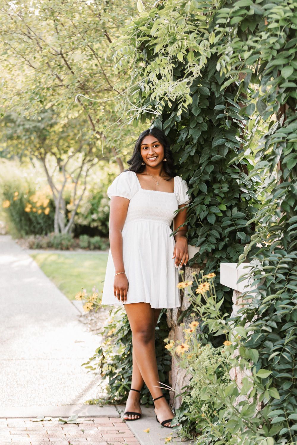 Full length portrait in white dress along garden path with greenery.