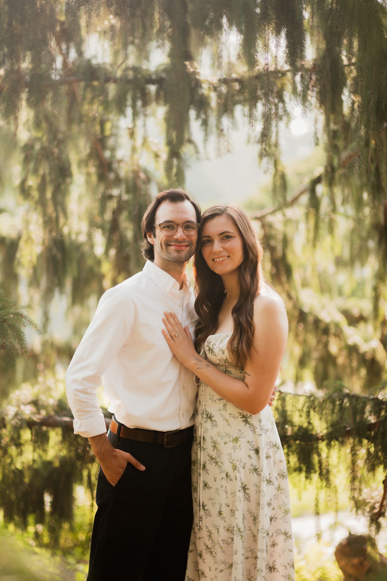 A smiling couple poses together outdoors beneath hanging moss-covered trees in warm golden light.