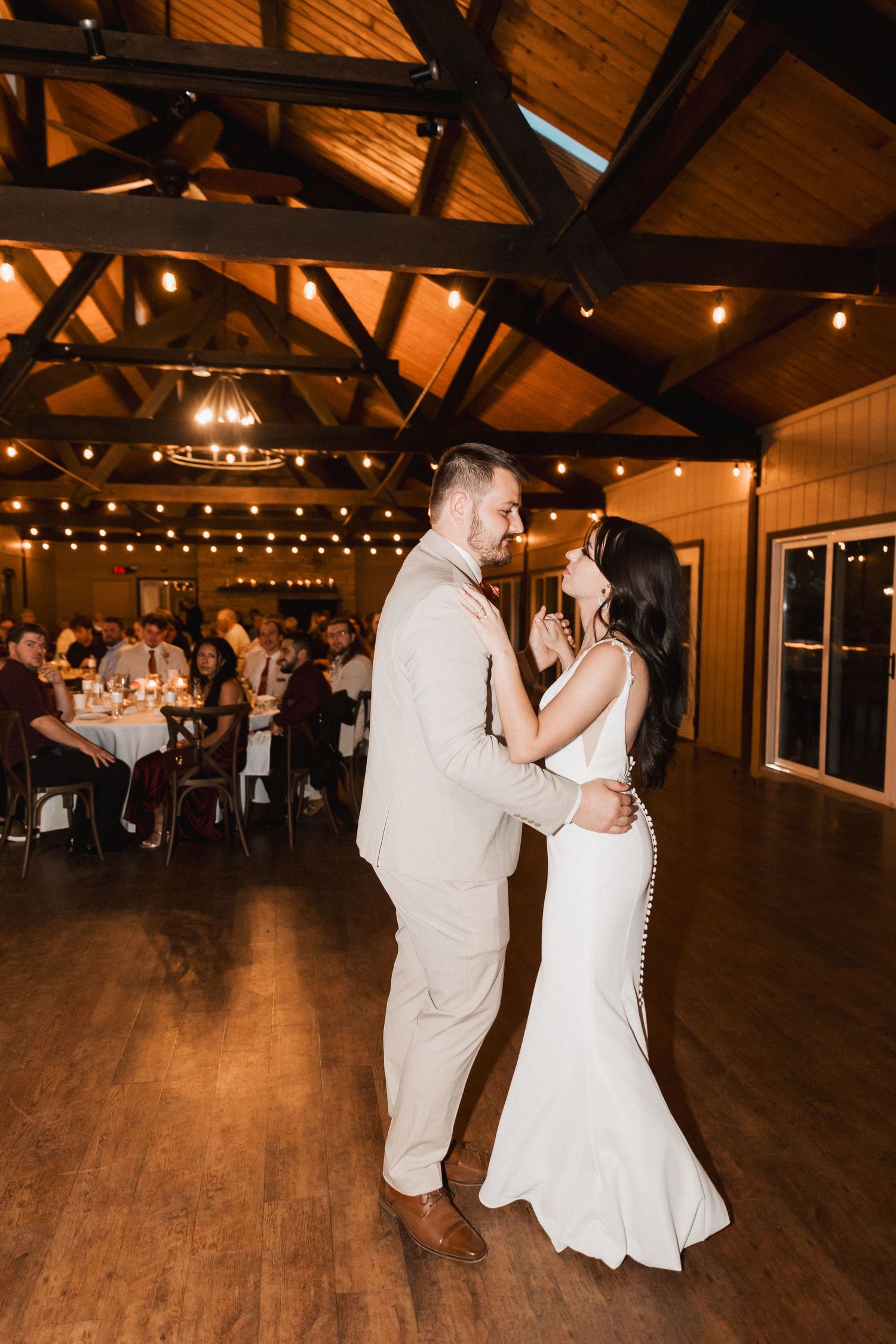 A couple in white attire dances together on a wooden floor in a rustic venue with string lights overhead.