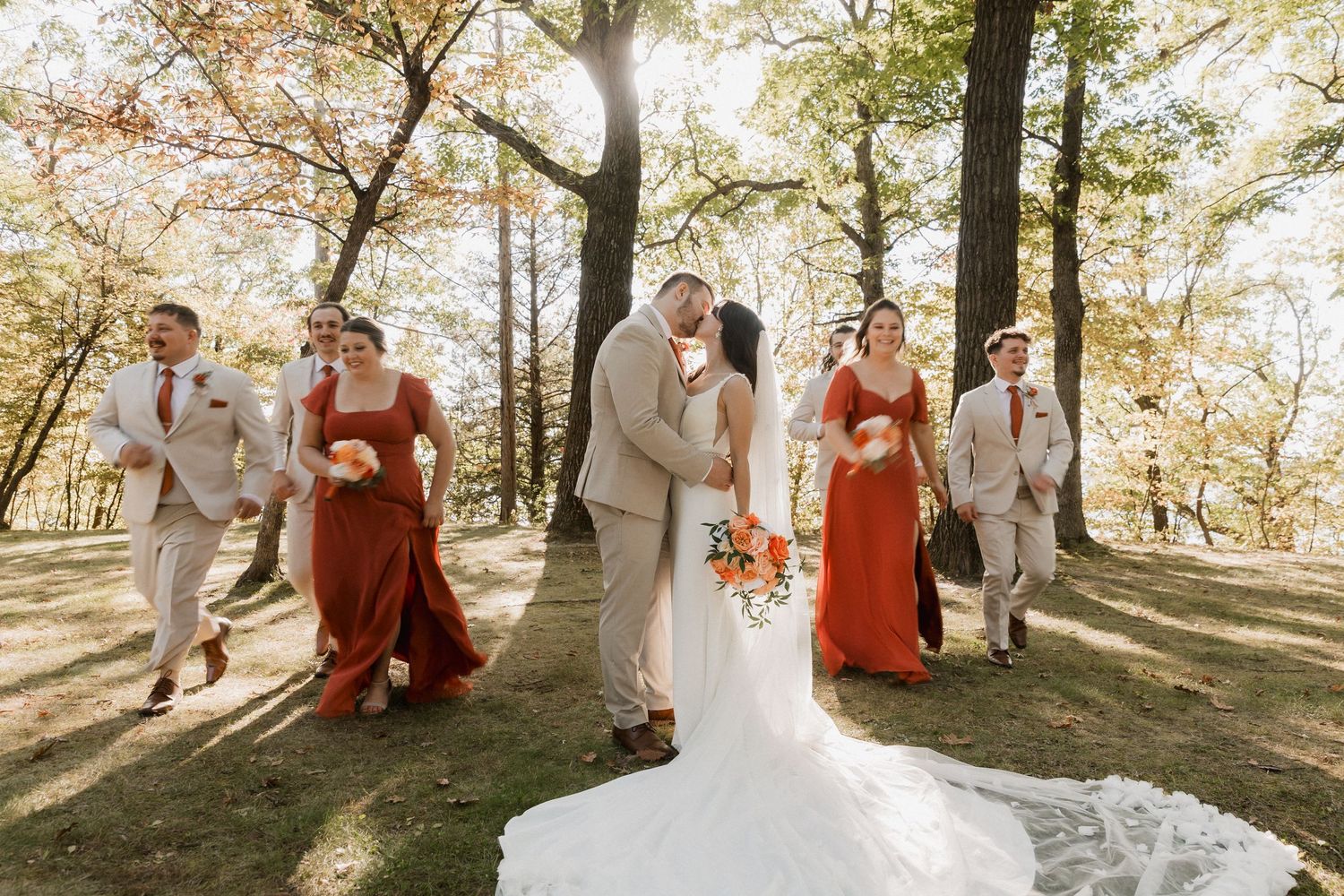 Wedding party walks outdoors in autumn, bride and groom kiss while bridesmaids in rust dresses and groomsmen in tan suits surround them.