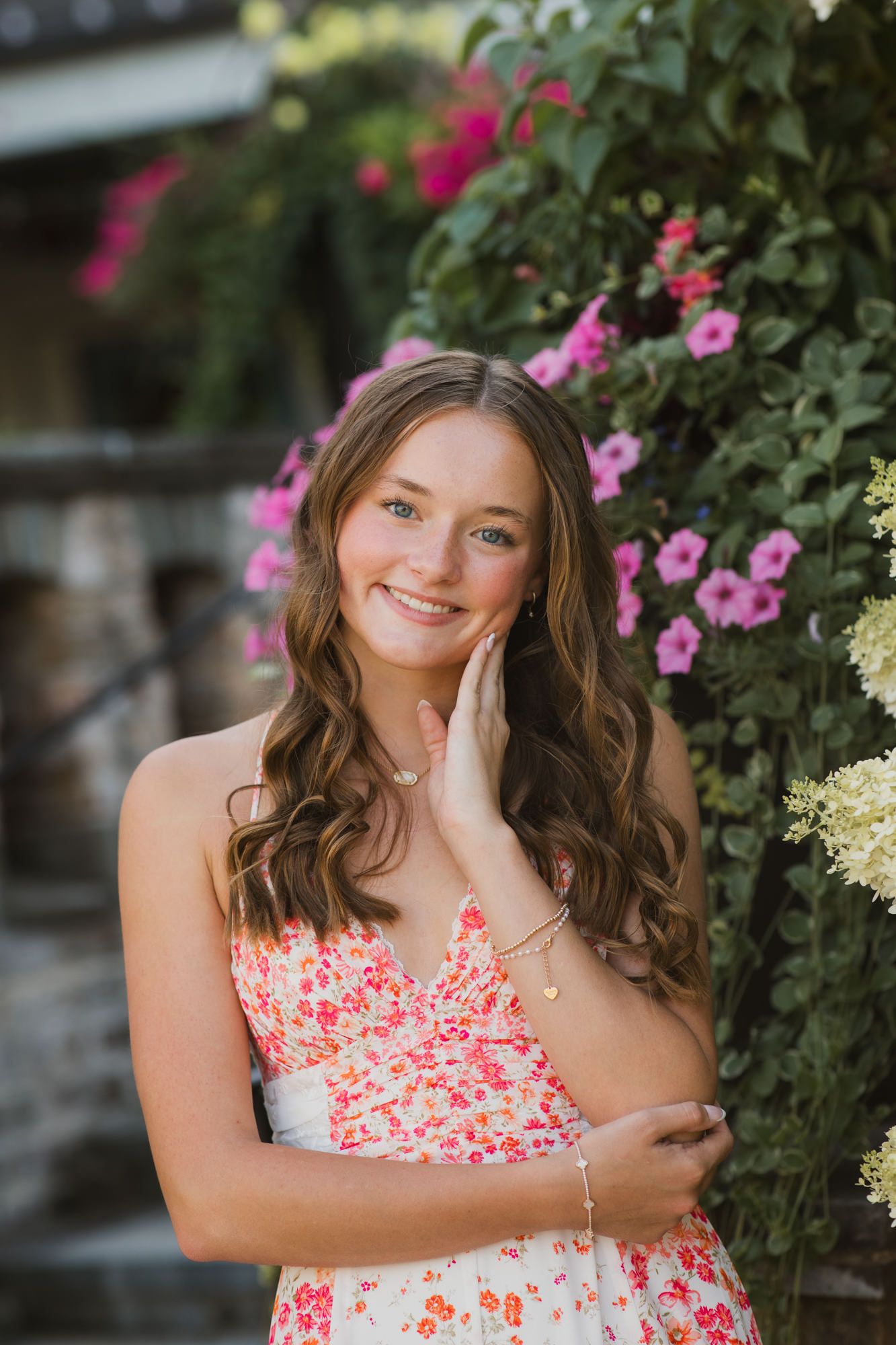 Young woman in floral dress smiling beside pink flowers and lush greenery in an outdoor garden setting.