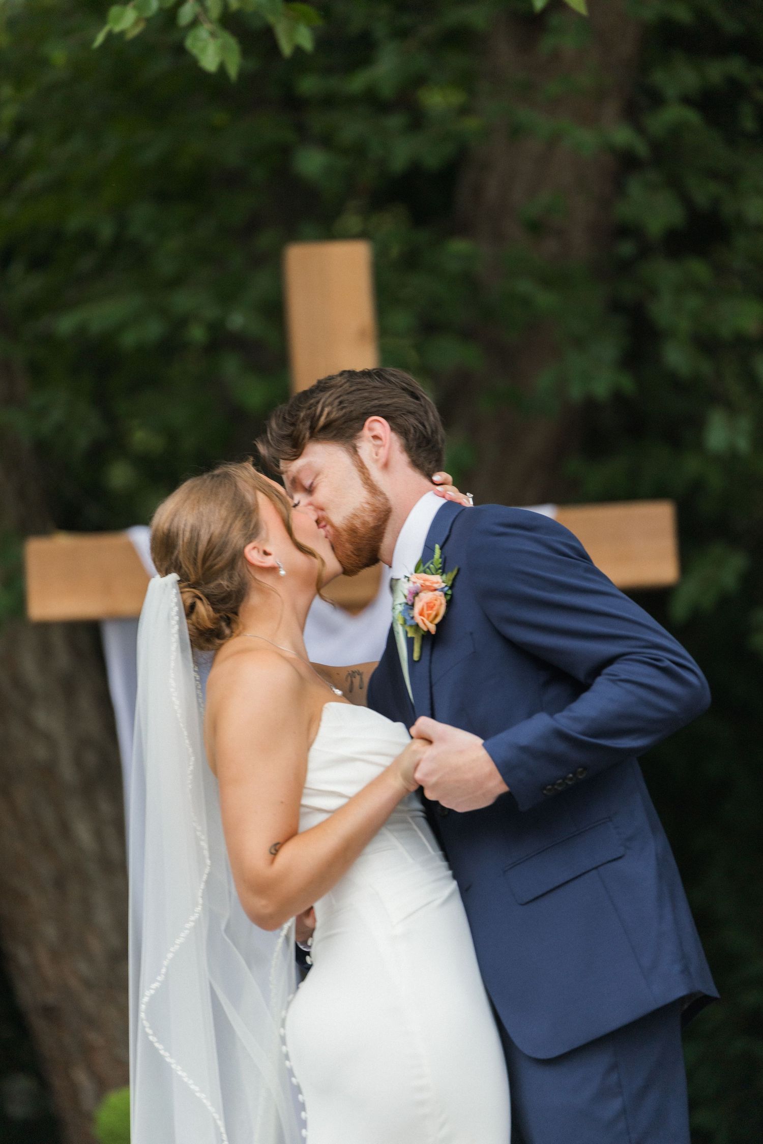 Bride and groom share first kiss in front of wooden cross at outdoor wedding.