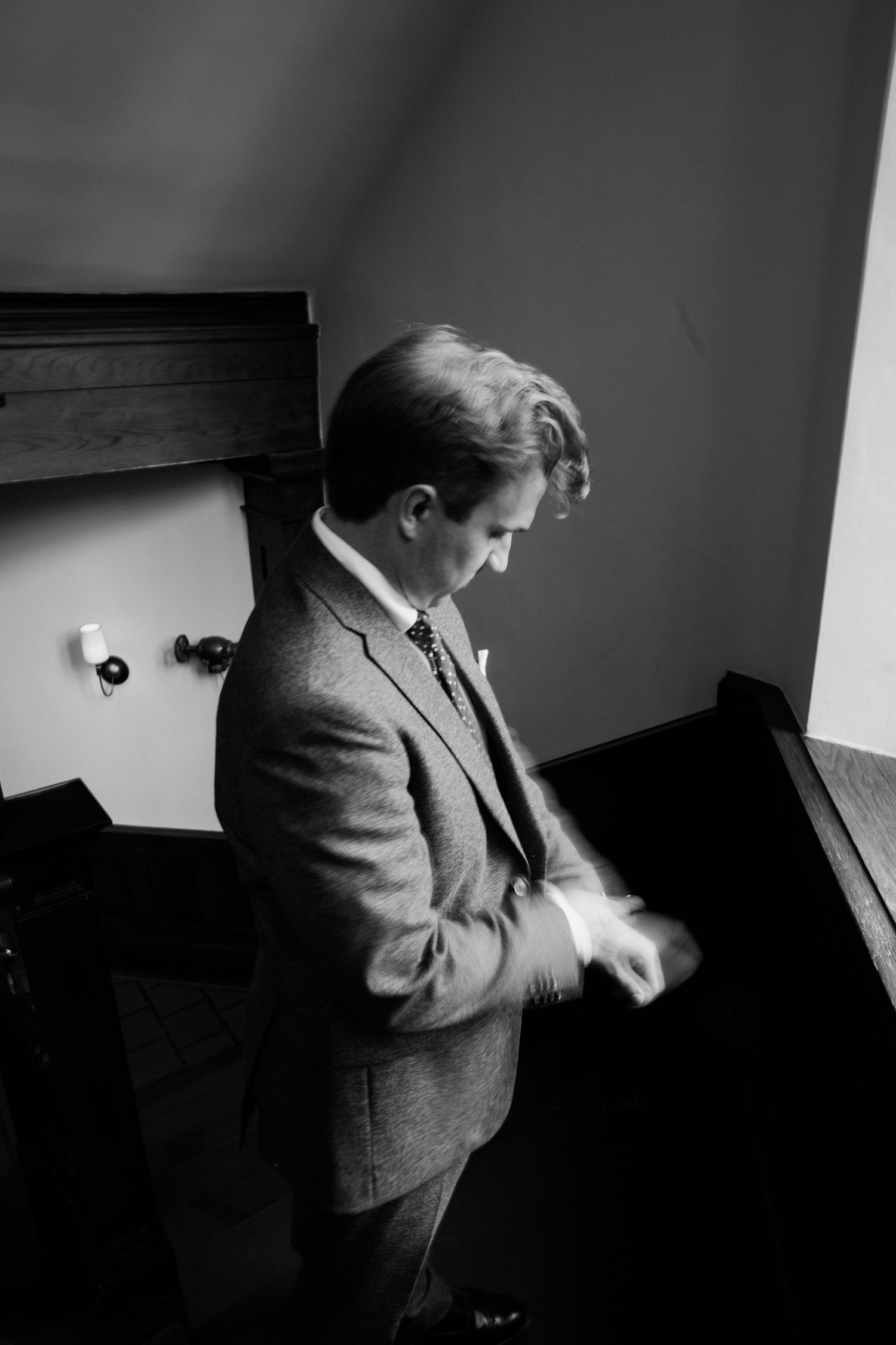 Groom in a gray suit adjusting his cufflinks in a dimly lit room, black and white wedding photo.