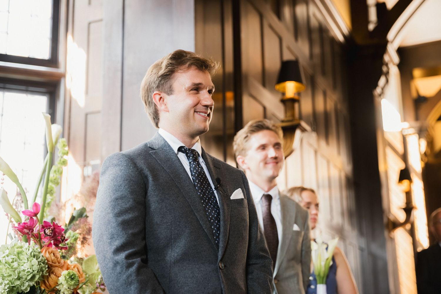 Groom smiling at the altar in a grey suit and dotted tie during an elegant indoor wedding ceremony.