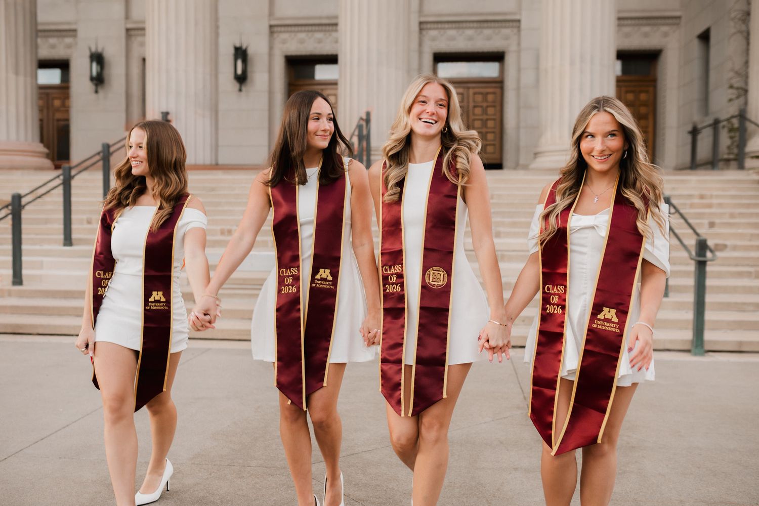 Four women in white dresses and maroon graduation stoles walk hand-in-hand on college campus steps.