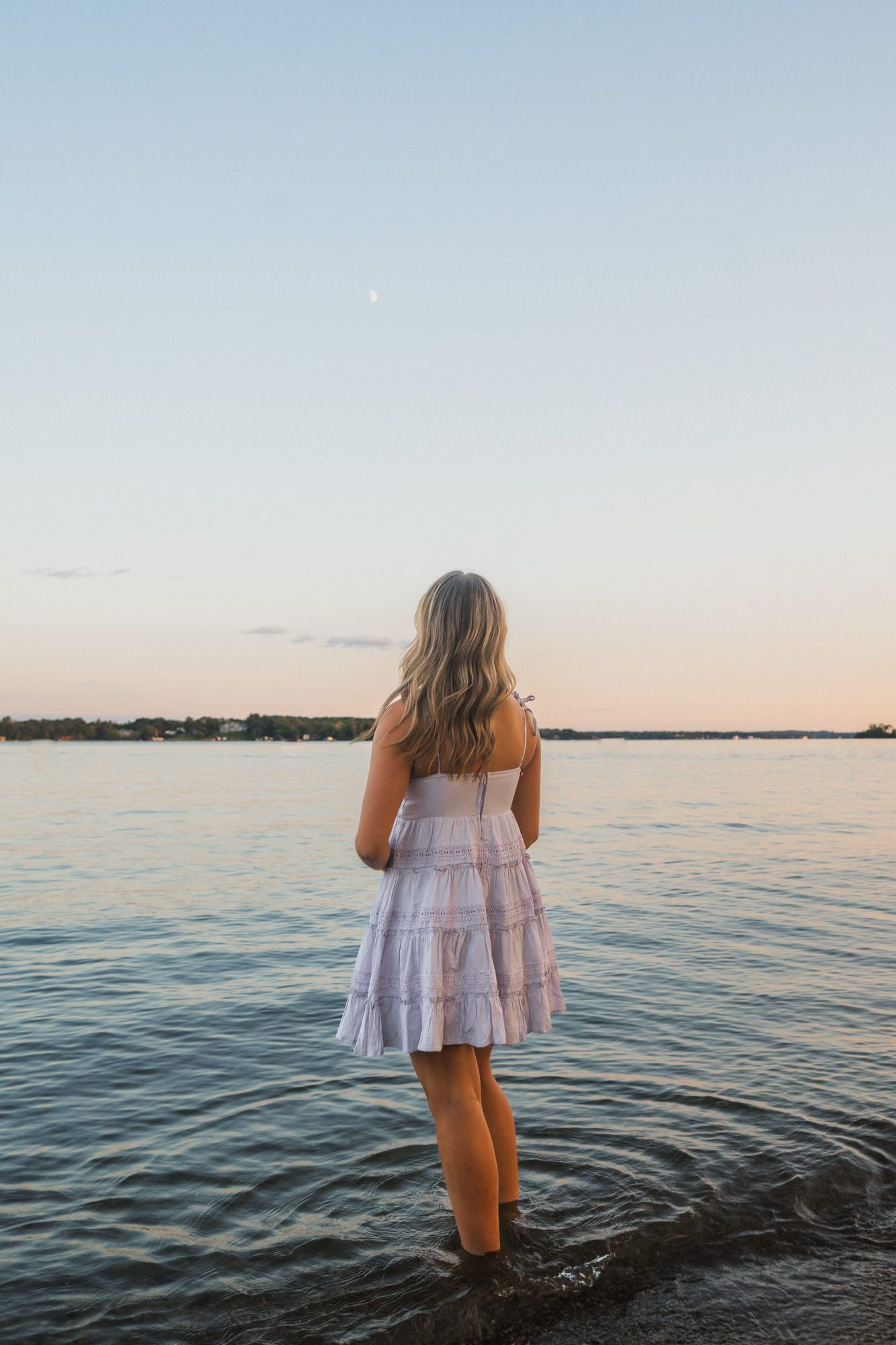 A blonde woman in a white sundress stands in shallow water at sunset, gazing at the calm horizon.
