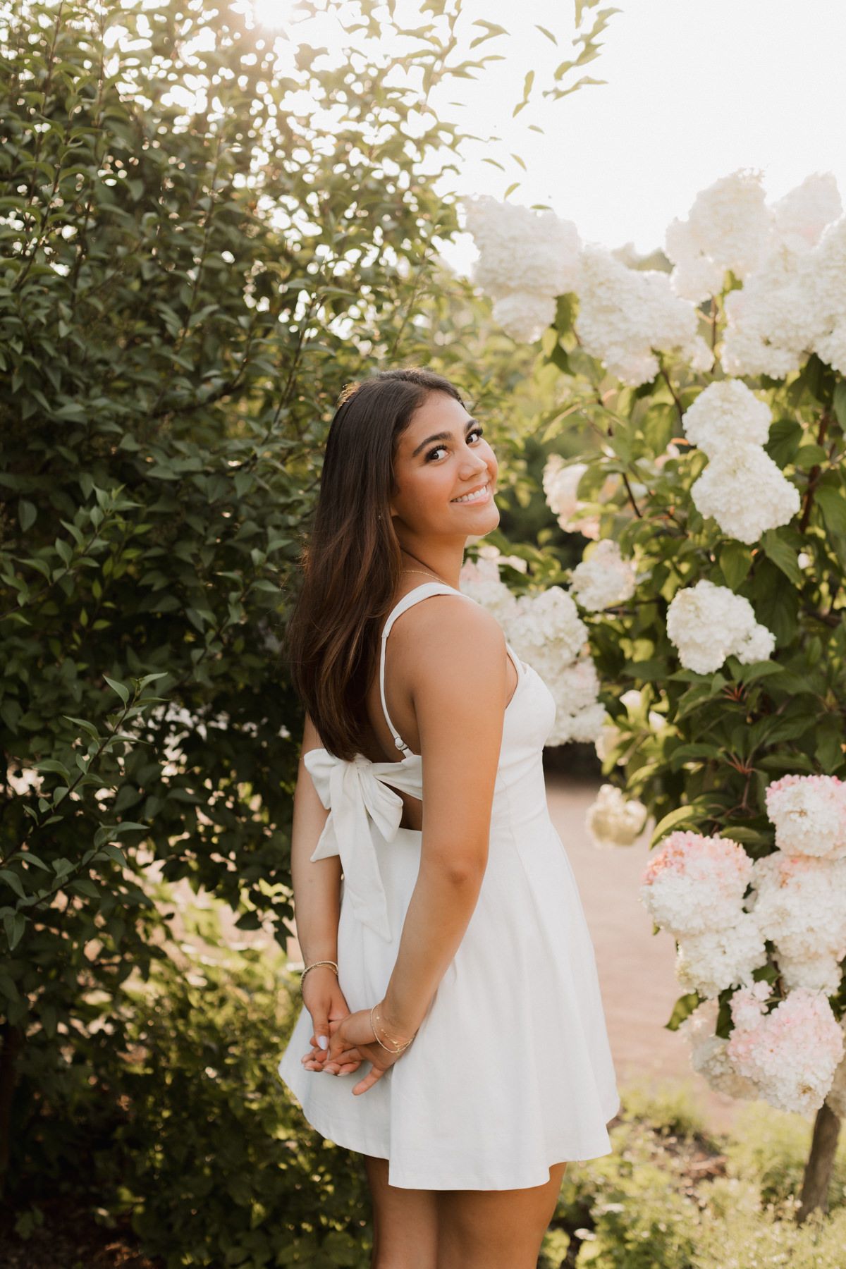 Young woman in white mini dress with bow back standing among white hydrangea blooms.