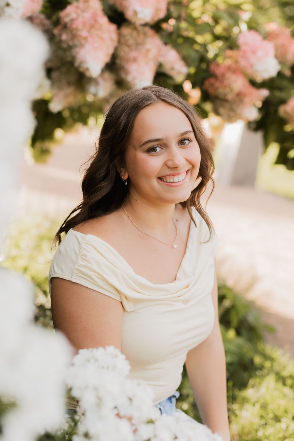 Smiling young woman in cream off-shoulder dress surrounded by pink and white hydrangeas outdoors.