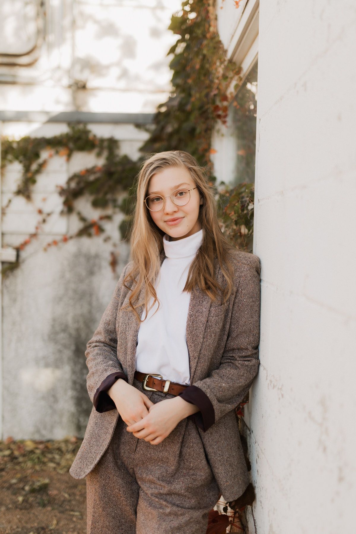 Young blonde woman in brown blazer suit and glasses leaning against white marble wall outdoors.