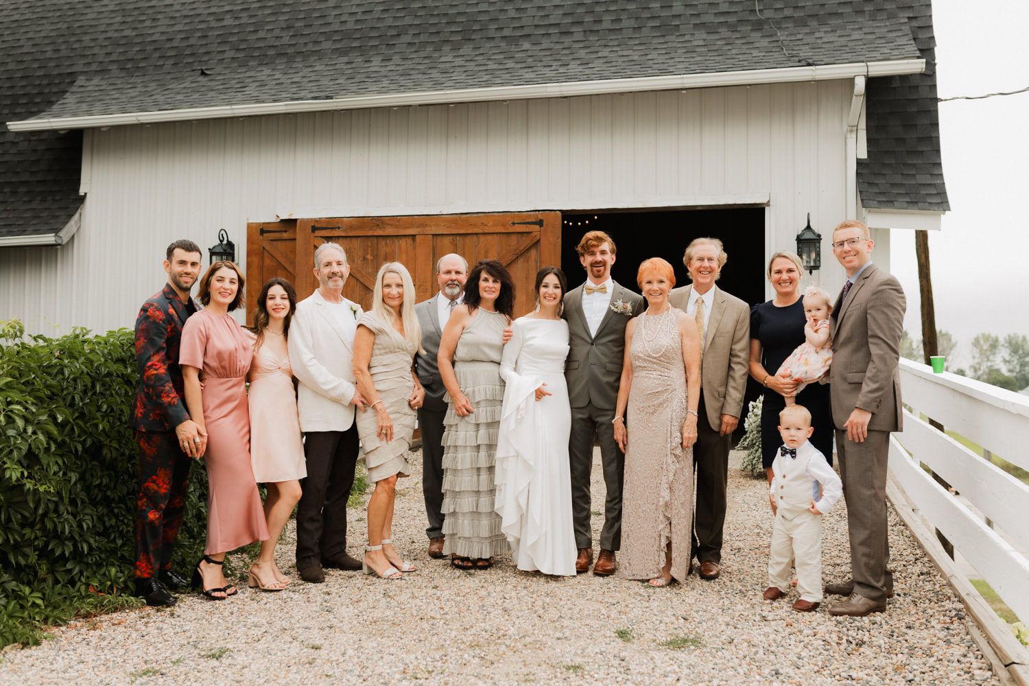 A wedding party poses together in front of a white barn wearing coordinated formal attire in neutral tones.