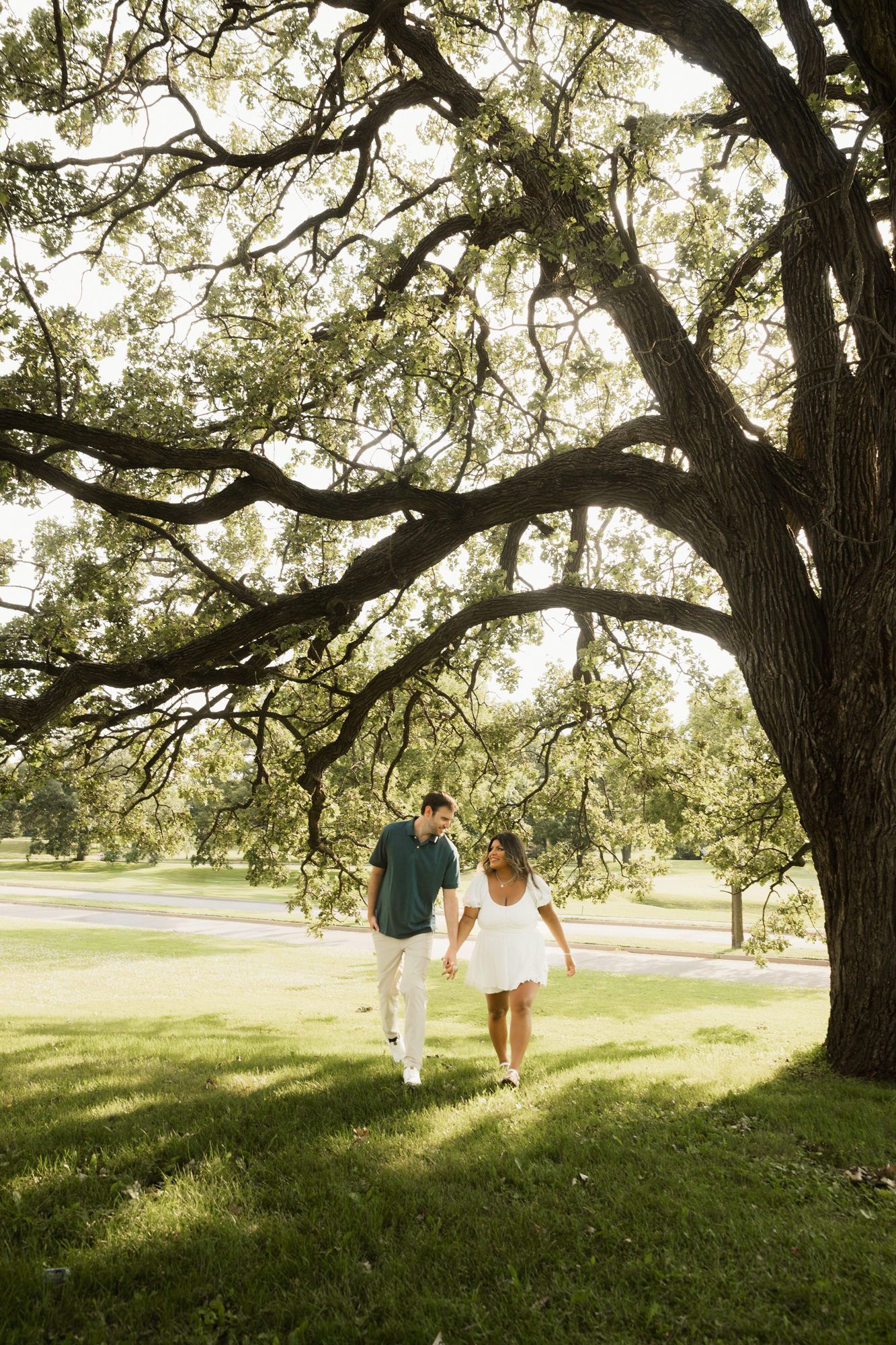 Couple holding hands walking under a large oak tree in a lush green park, woman in white dress, man in teal shirt.
