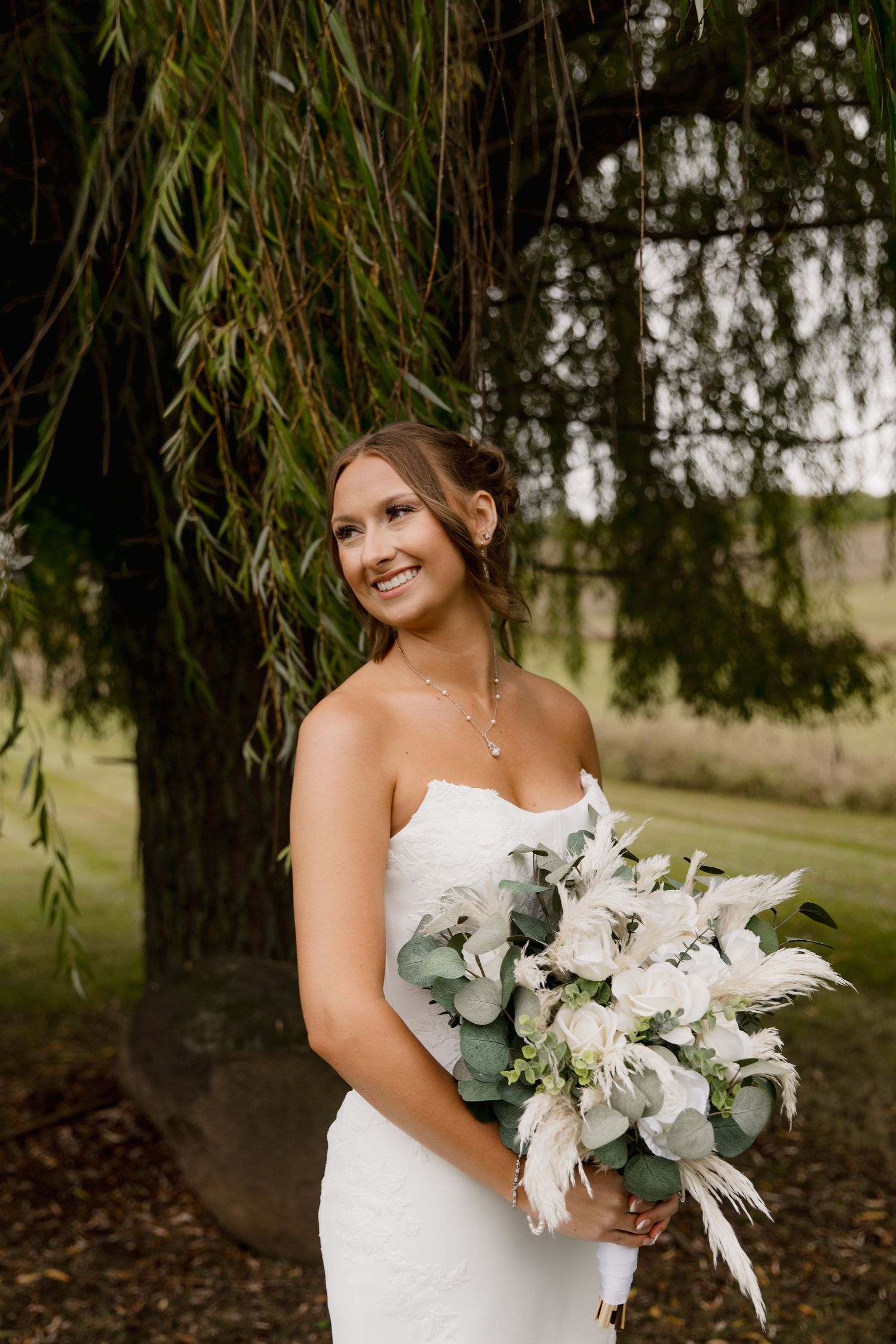 Smiling bride in strapless white gown holds white floral bouquet with eucalyptus under a weeping willow tree.