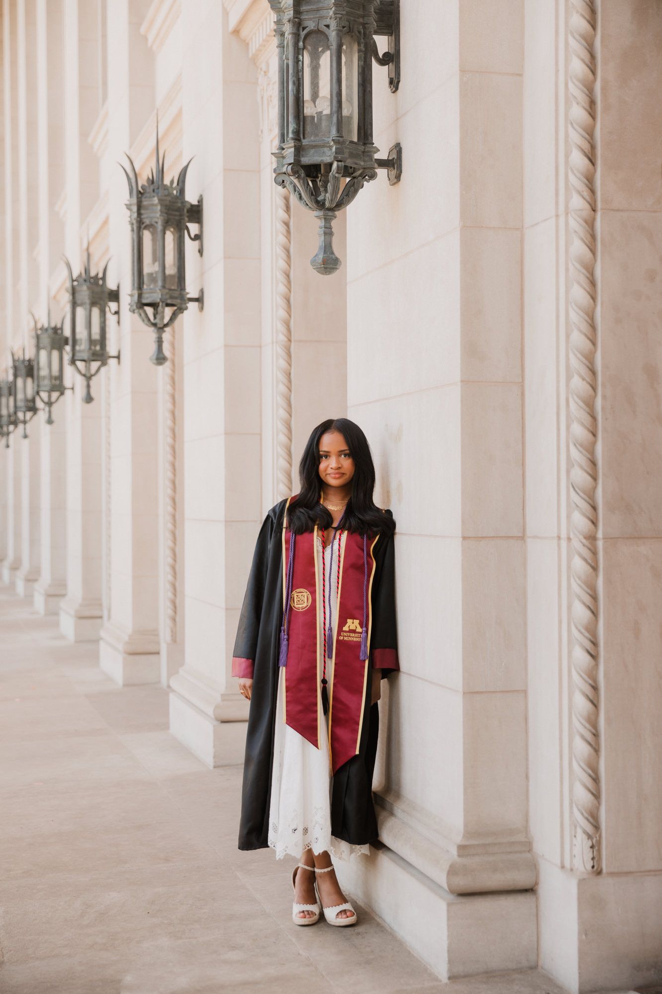 Graduate in black gown and maroon University stole leans against stone column in classic building hallway.