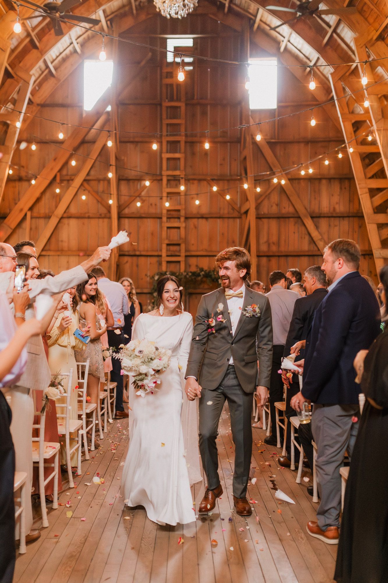 Bride and groom walk down barn wedding aisle amid cheering guests, string lights, and rustic wood architecture.