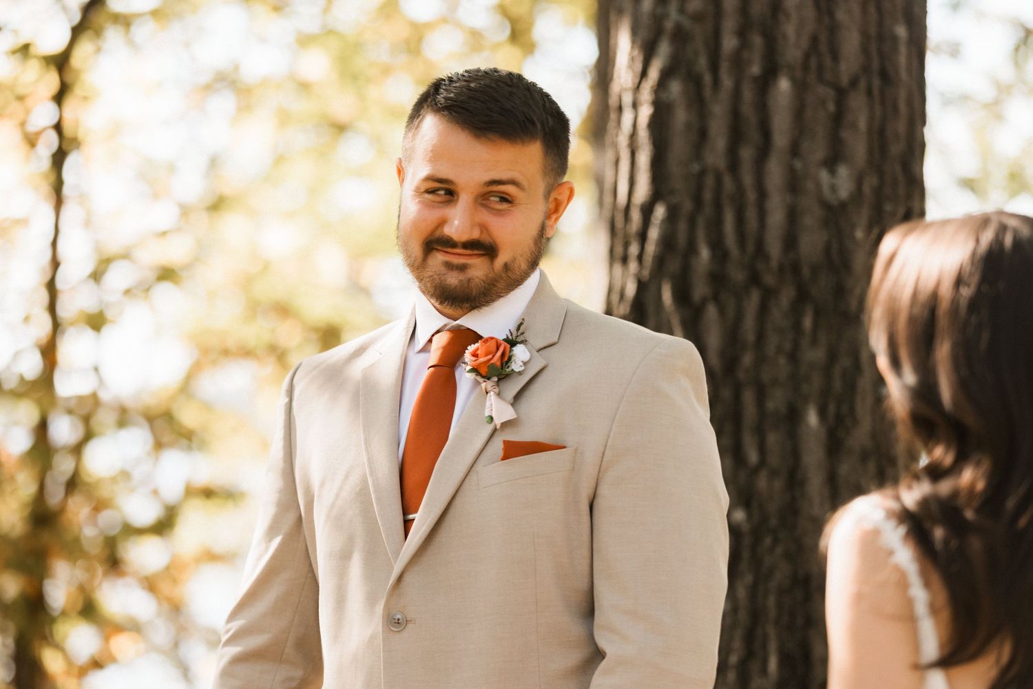 A couple in formal wedding attire embraces outdoors with warm sunlight filtering through trees in the background.