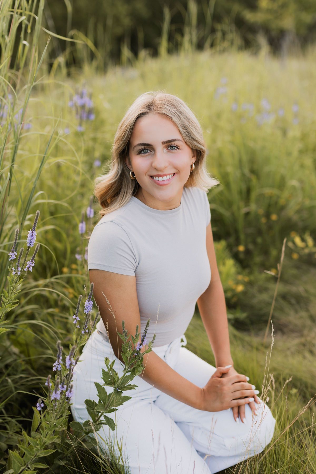 Young blonde woman smiling in a wildflower meadow, wearing a grey top and white skirt.