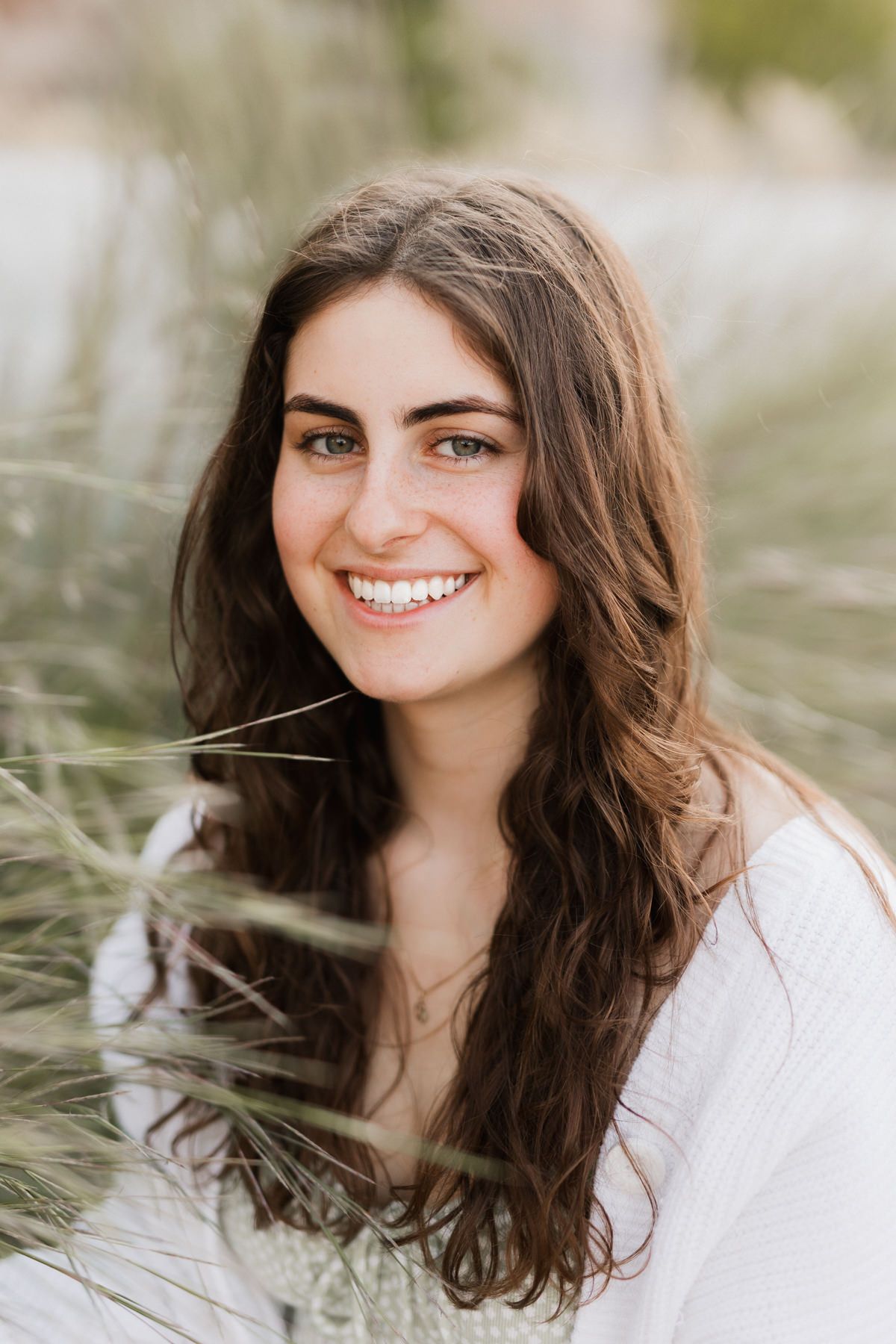 Young woman with long brown hair smiling outdoors among tall grass in a white top.