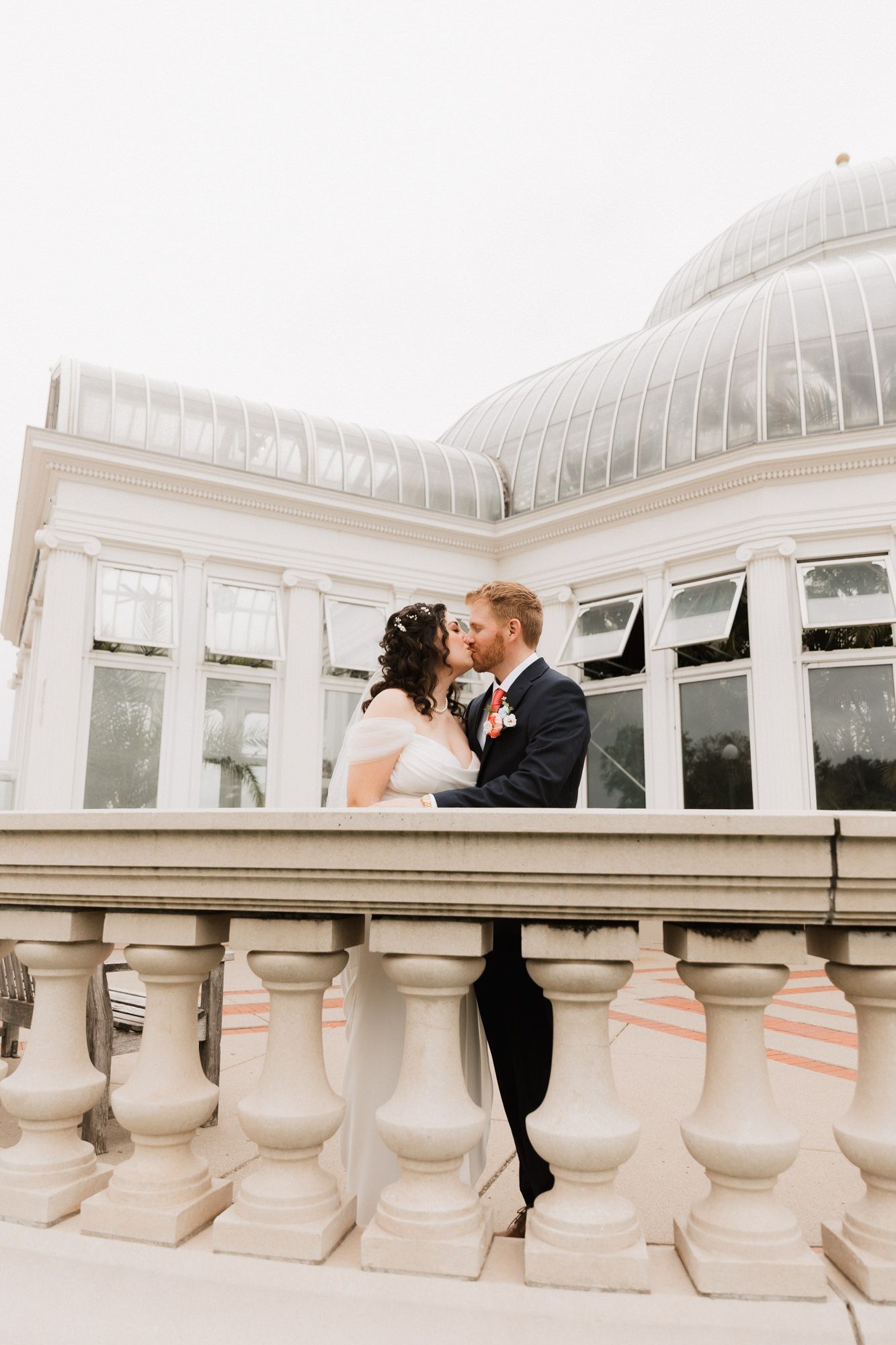 A bride and groom share a romantic kiss on a balustrade balcony in front of an elegant glass conservatory.