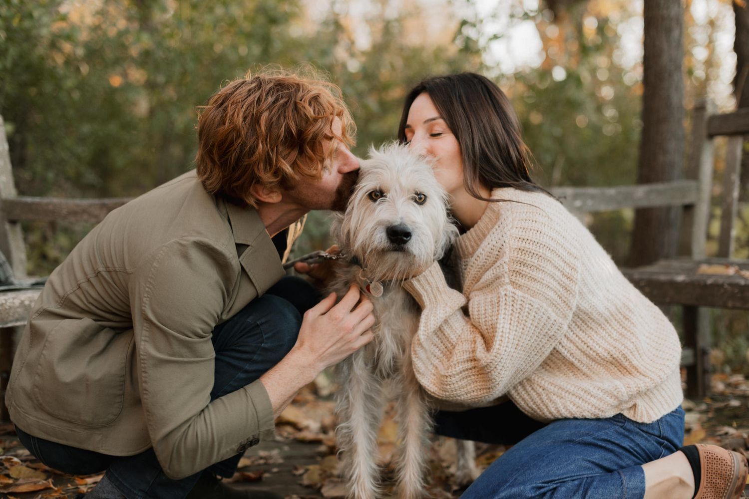 Couple kissing their fluffy white dog outdoors in an autumn forest setting.
