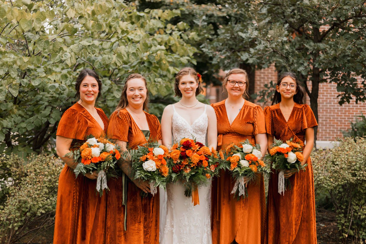 Bridal party in burnt orange bridesmaid dresses poses outdoors with bouquets against a green garden backdrop.