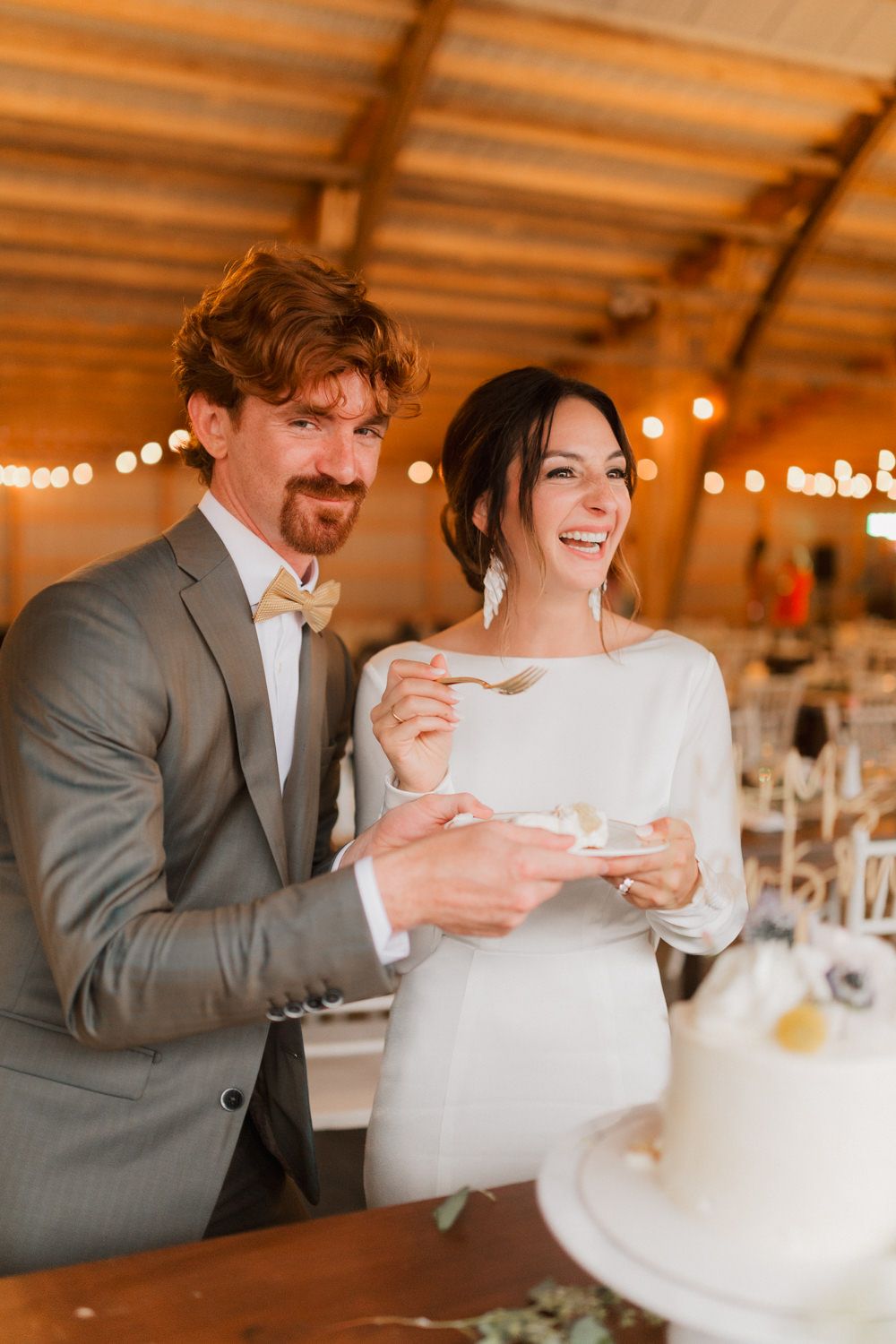 A couple shares a joyful moment while cutting their wedding cake in an indoor venue with string lights.