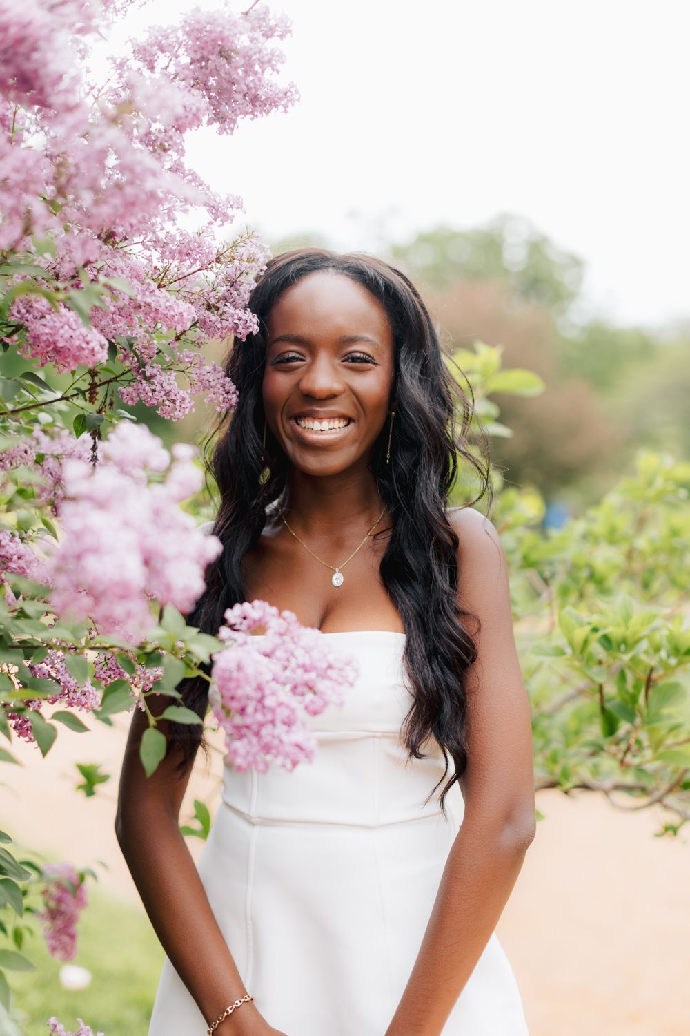 Graduate in white dress posing among blooming pink cherry blossoms.