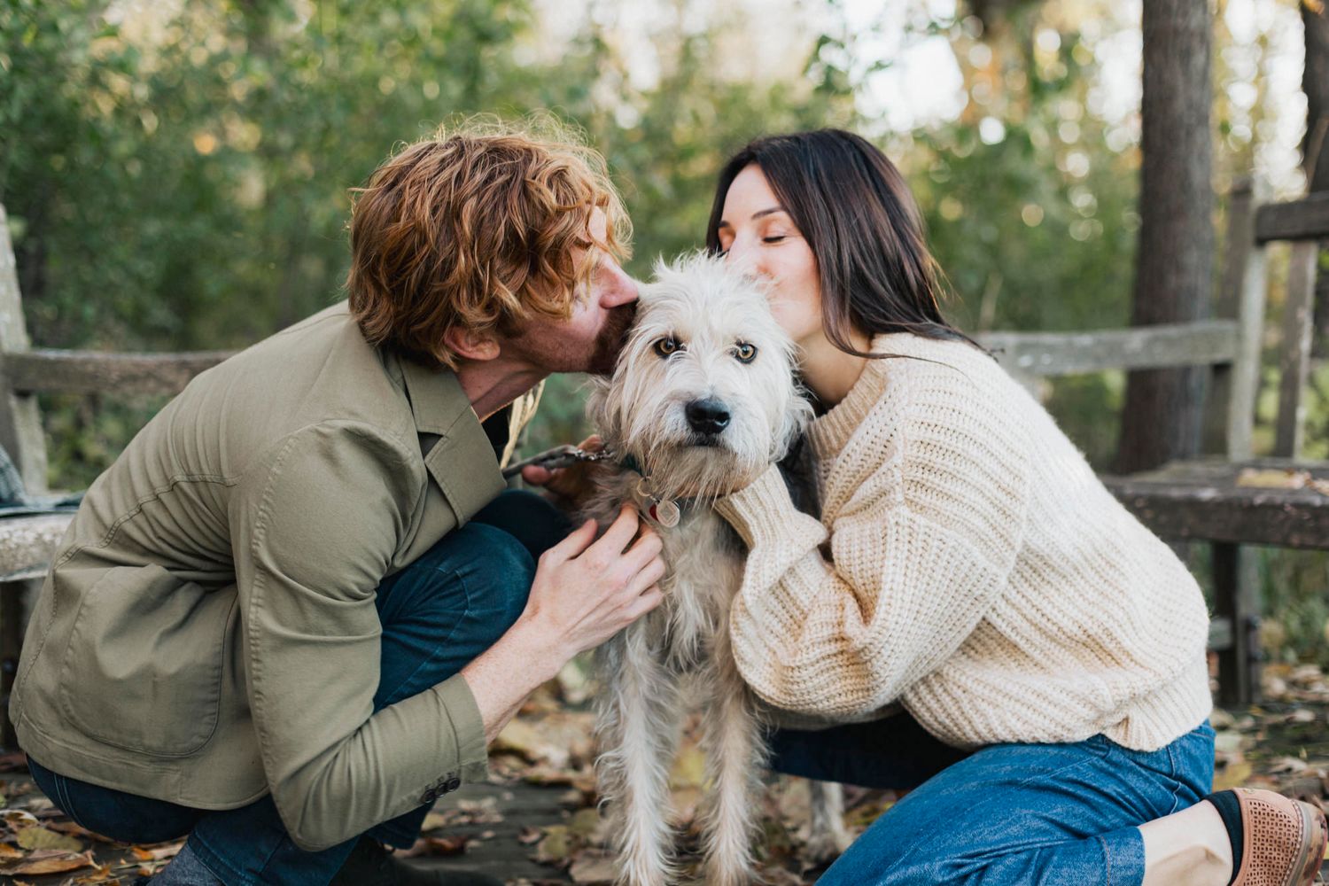A couple kneels down to pet and kiss their fluffy white dog on a wooden deck.