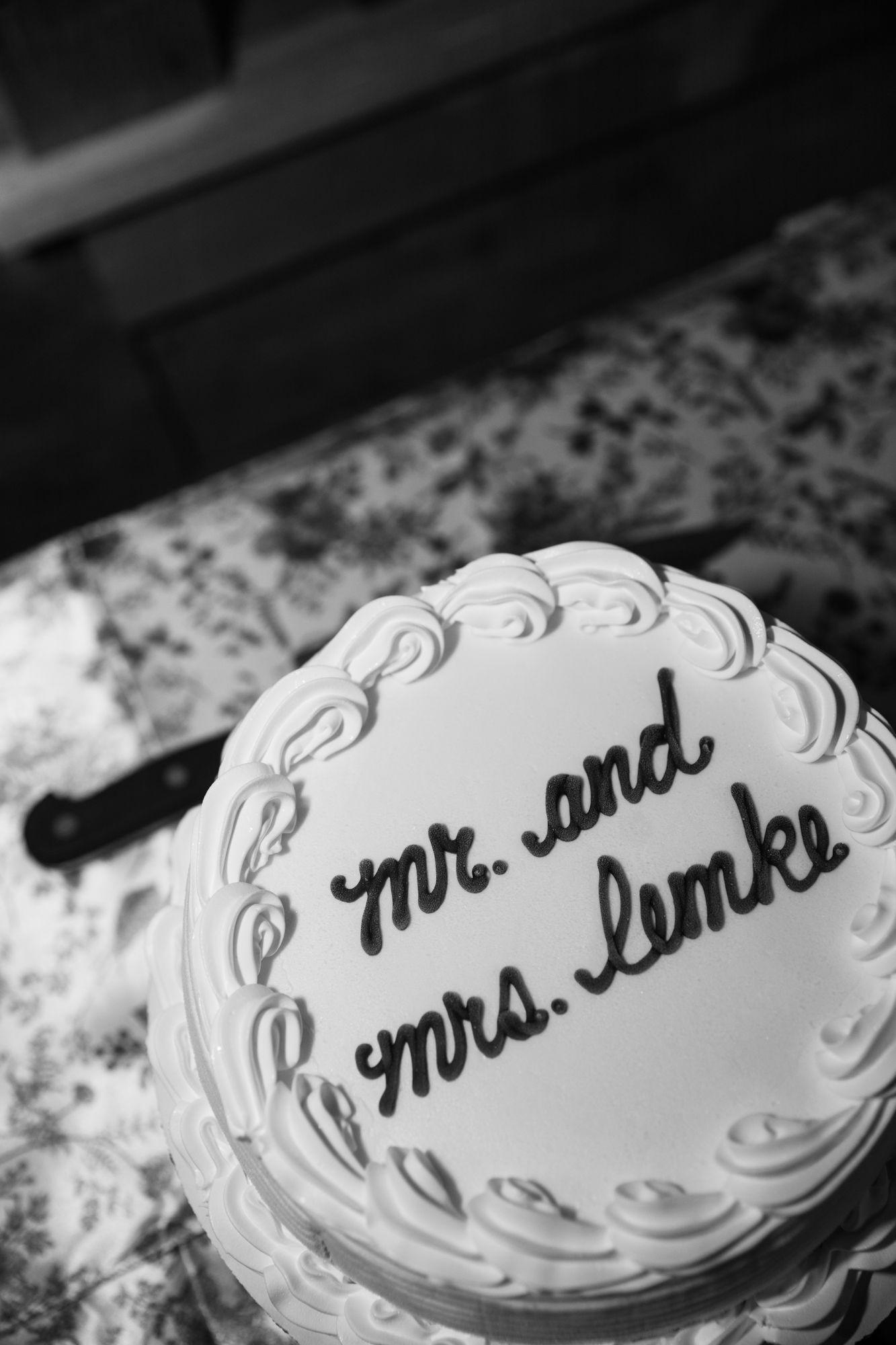 Black and white photo of a round wedding cake decorated with frosting and Mr. and Mrs. Lemke written in cursive.