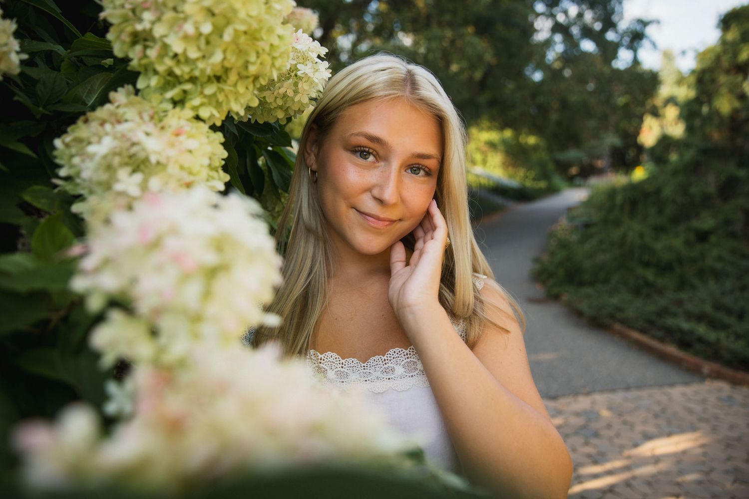 Young blonde woman posing among white hydrangea flowers in a sunlit garden path, wearing a white dress.