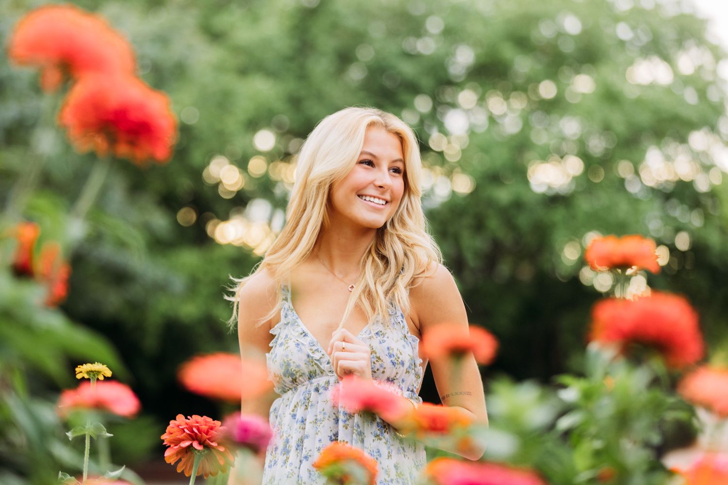 A woman in a floral sundress poses in a garden filled with vibrant orange and pink zinnias during sunset.