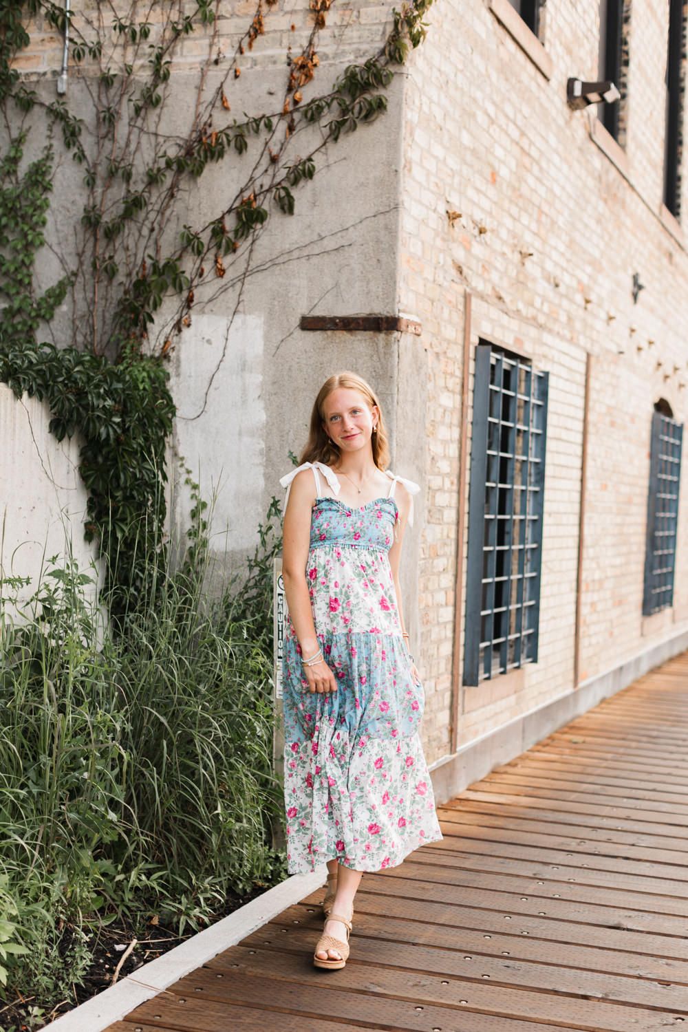 A summery floral maxi dress is worn on a wooden deck walkway next to a brick building with climbing vines.