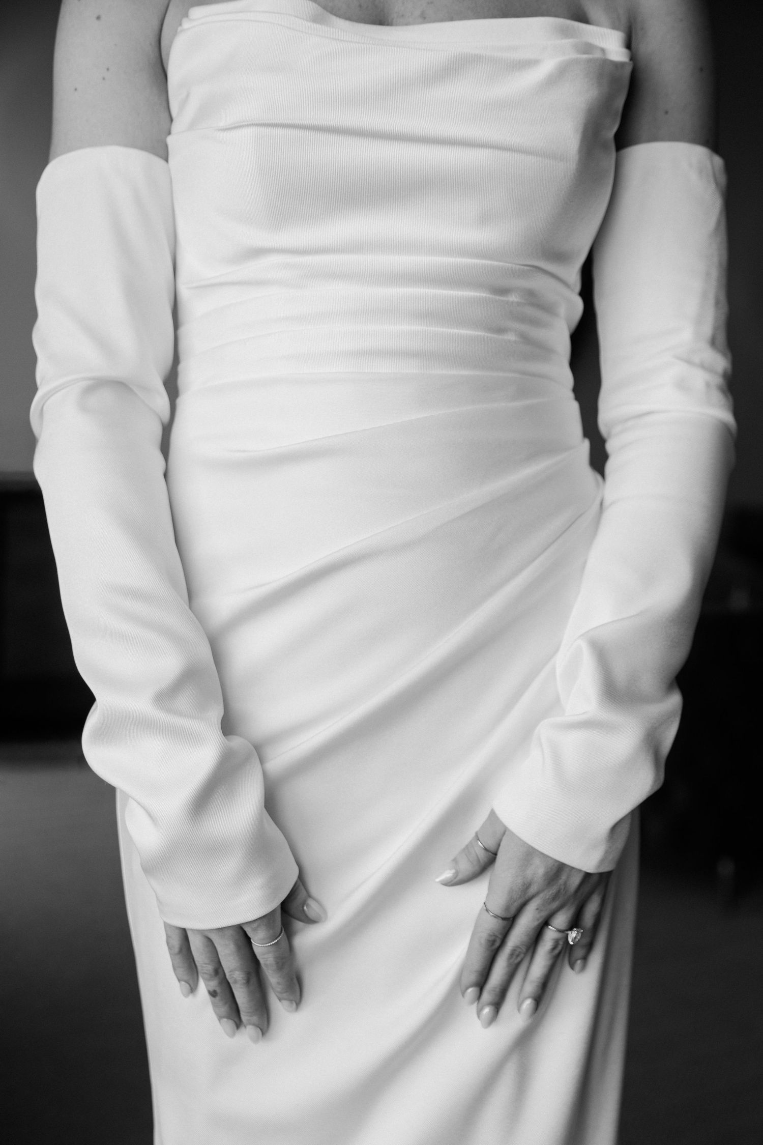 Black and white close-up of a bride's satin gown with long sleeves, her hands resting at her sides.