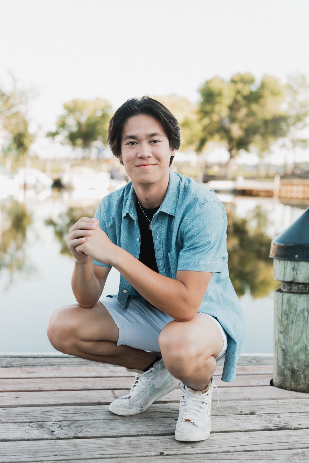 A person in a light blue shirt crouches on a wooden dock by a serene lake on a sunny day.