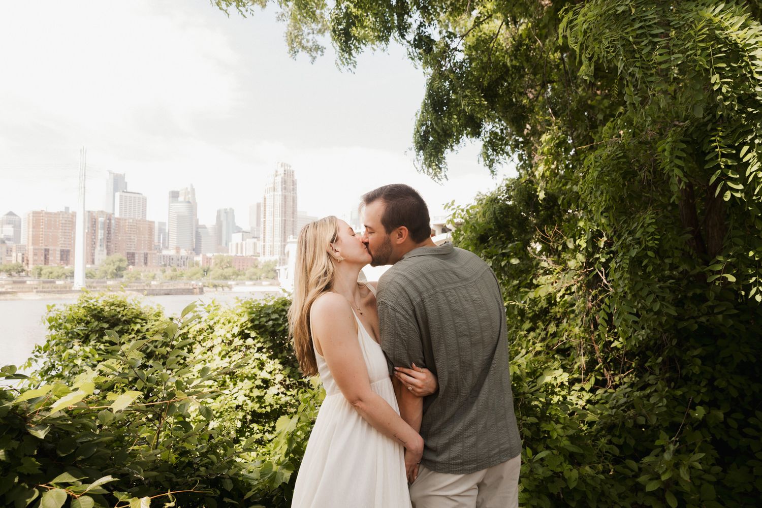 Couple kissing in lush green garden with city skyline and river in background on sunny day.