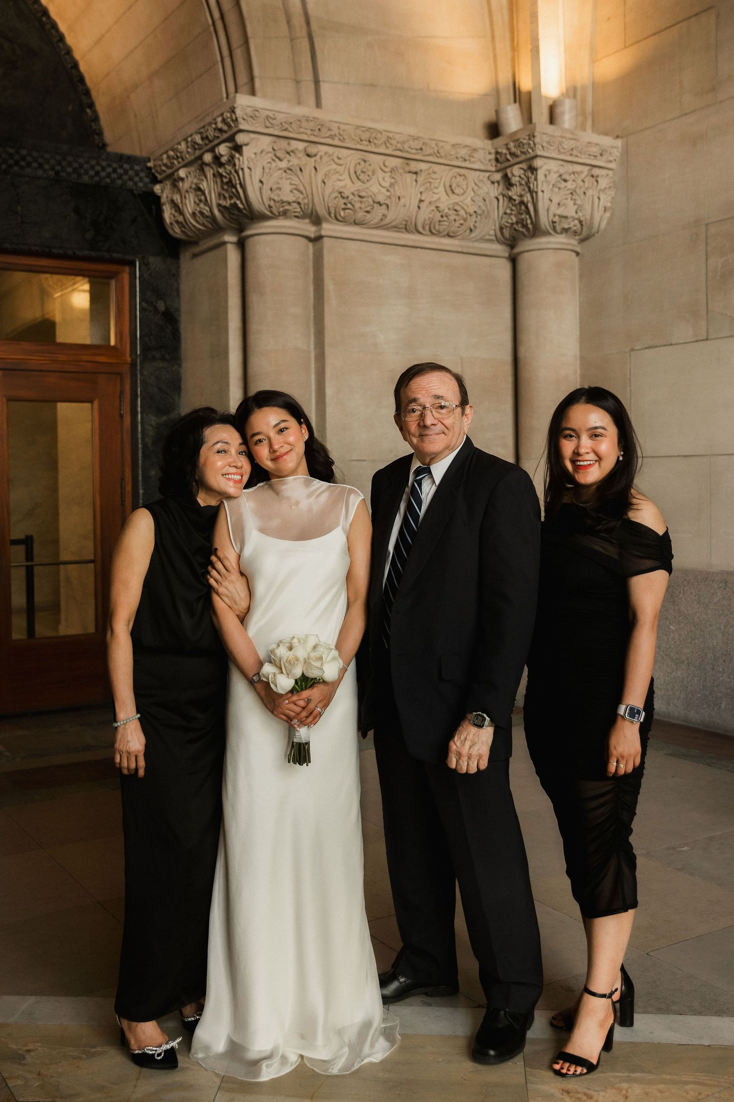 Bride in white gown holds bouquet with family in black attire posing inside elegant stone building.