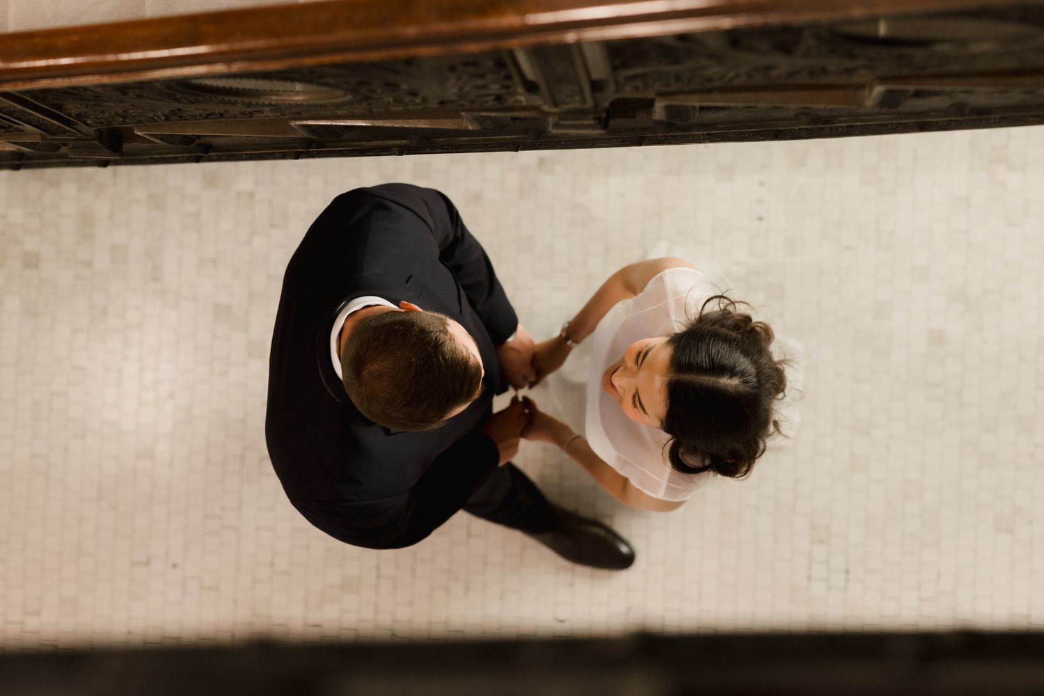 Aerial view of two people shaking hands on a tiled floor, one in a dark suit and one in a white shirt.