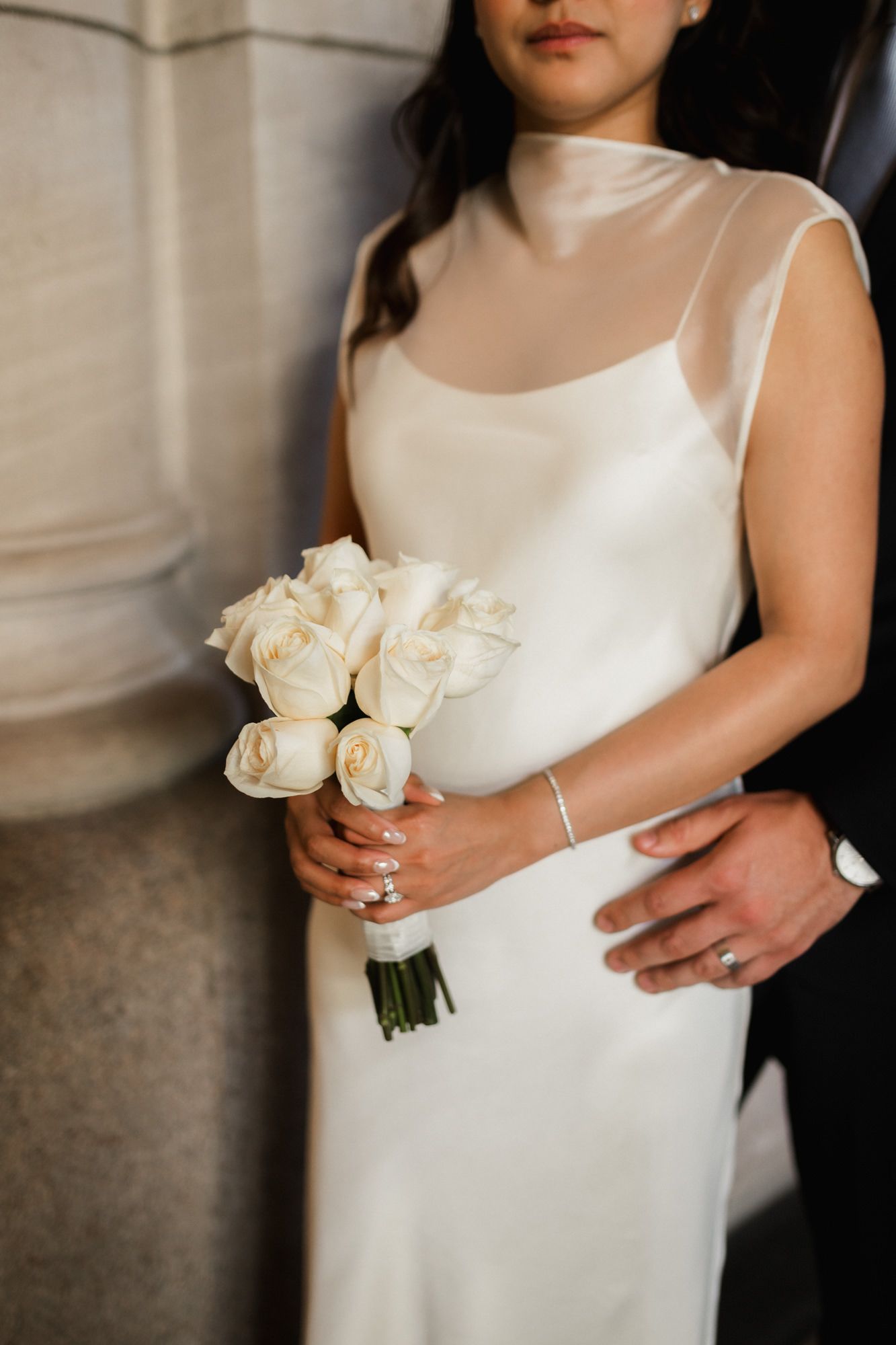 Bride in white satin gown with sheer neckline holds white rose bouquet while groom places hand on her waist.