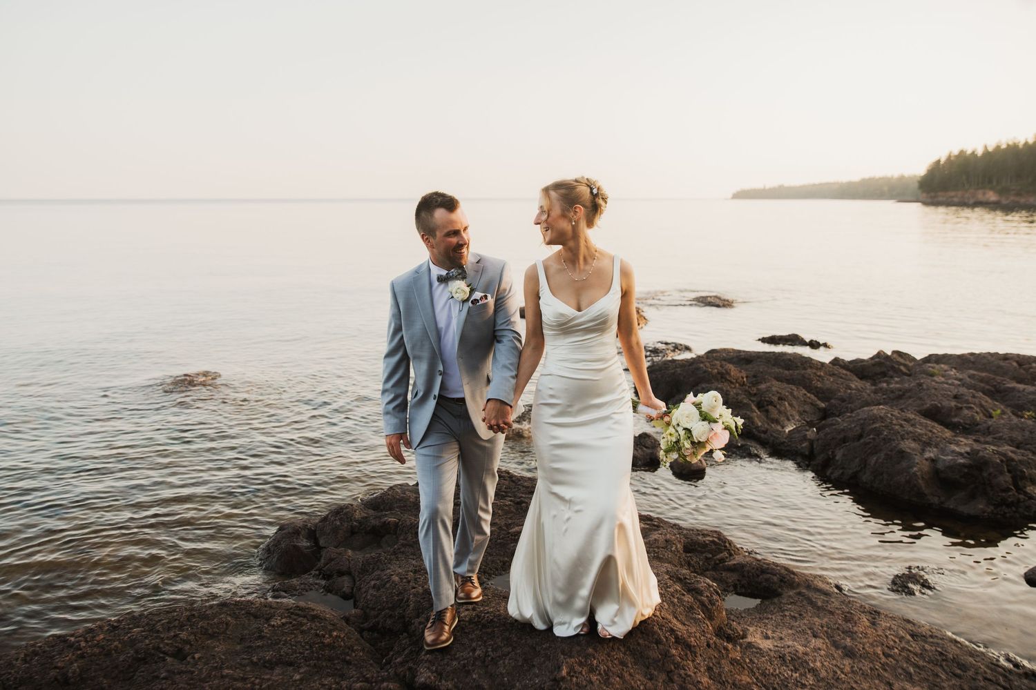 Bride and groom holding hands on rocky lakeside shore at sunset, bride in white gown holding bouquet, groom in light blue suit.